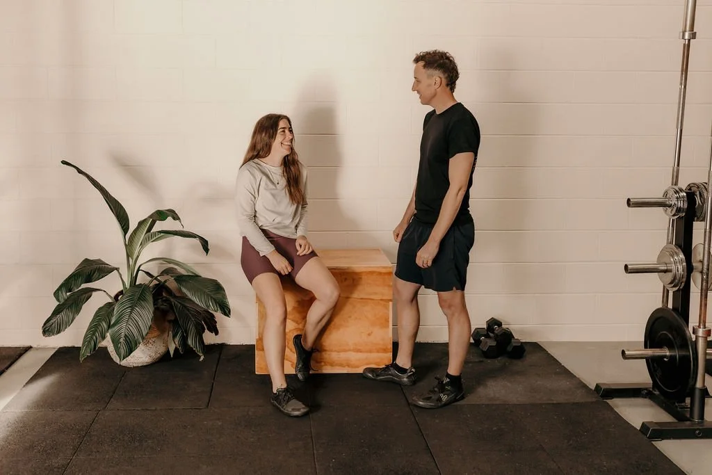 A man and woman with athletic attire are at a gym, talking and smiling. The woman is sitting on a wooden box, and the man is standing facing her. Gym equipment and a large leafy plant are in the background. 4b Gym Pambula NSW.