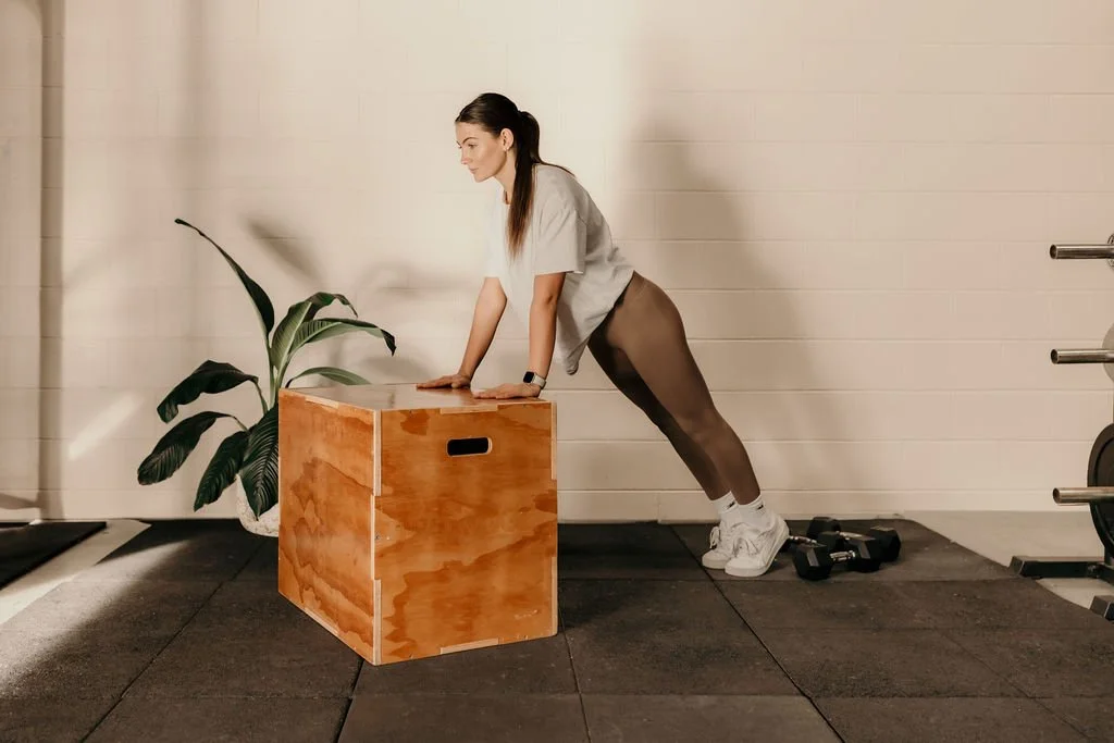 A woman performing a push-up exercise on a wooden box in a gym, with a potted plant nearby and dumbbells on the floor.