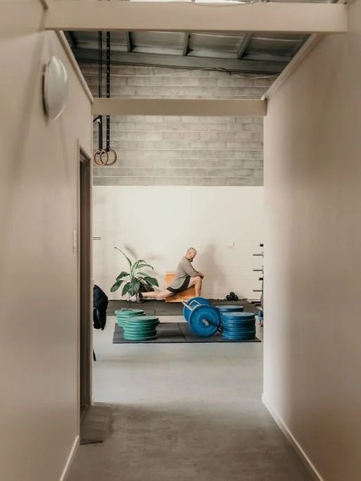 Person practicing yoga on a mat in a gym or fitness studio with weight plates stacked nearby, viewed through an indoor hallway. 4b Gym Pambula NSW.