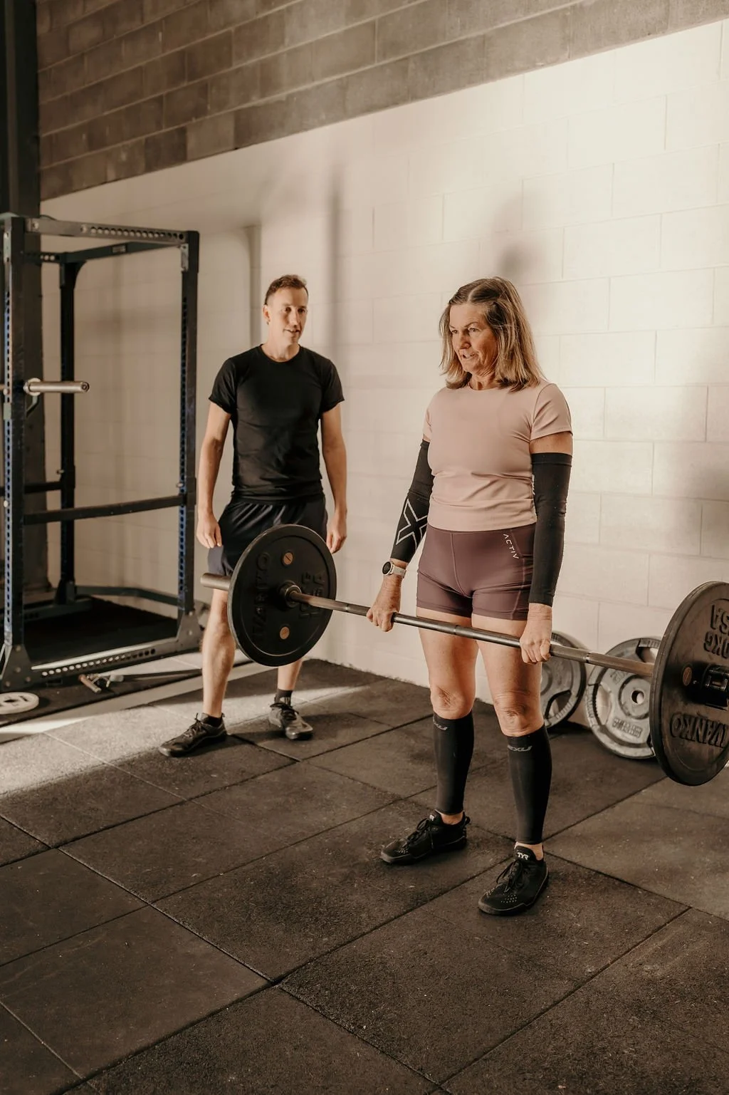 A woman in workout clothes lifting a barbell with weights at a gym, with a trainer standing nearby. 4b Gym Pambula NSW.