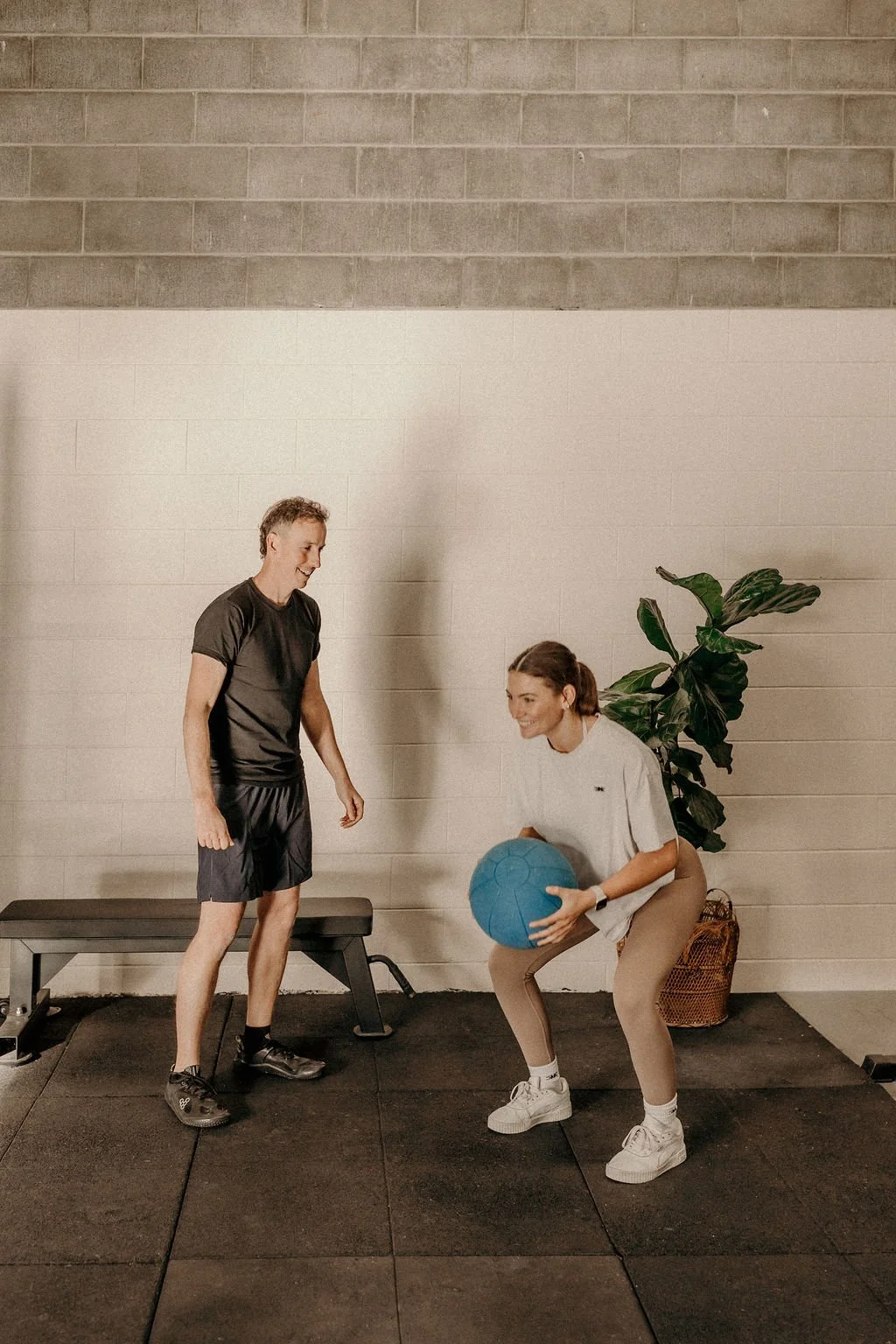 A man and a woman are in a gym, engaging in a workout with a blue medicine ball. The woman is in a squat position, holding the ball with both hands, while the man stands nearby, offering coaching support. 4b Gym Pambula NSW.