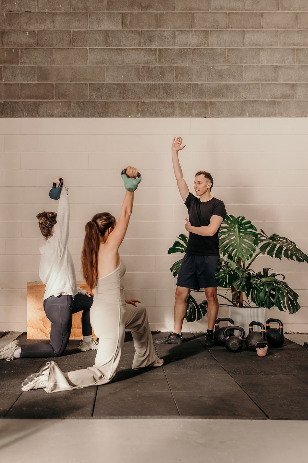 A personal trainer, Kean Lawler, instructs two women in a fitness class using kettlebells, with a large plant and kettlebells on the floor. 4b Gym Pambula NSW.