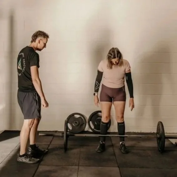 A woman preparing to lift a barbell while a man watches in a gym.