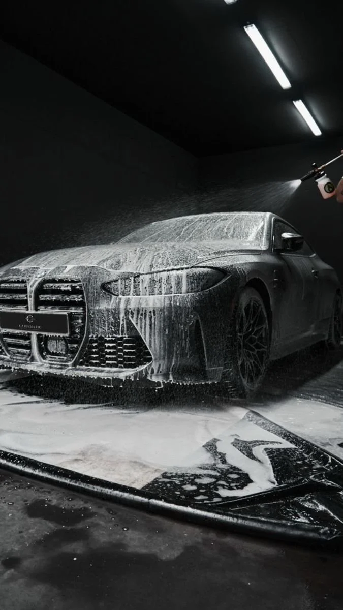 A dark-colored sports car being washed with soap and water in an indoor car wash bay, with bright overhead lights illuminating the scene.