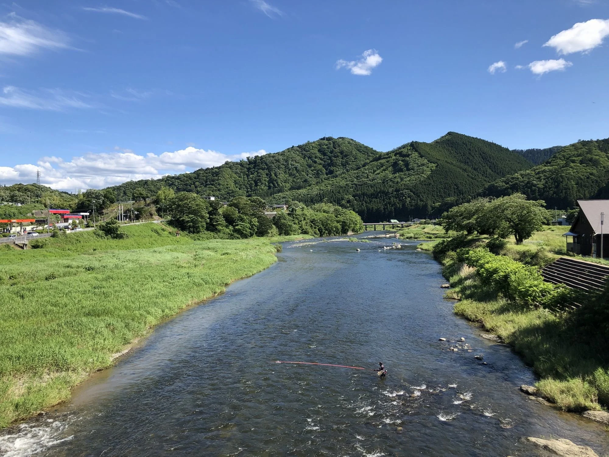 A river flowing through lush green grass with trees and mountains in the background, under a partly cloudy blue sky.