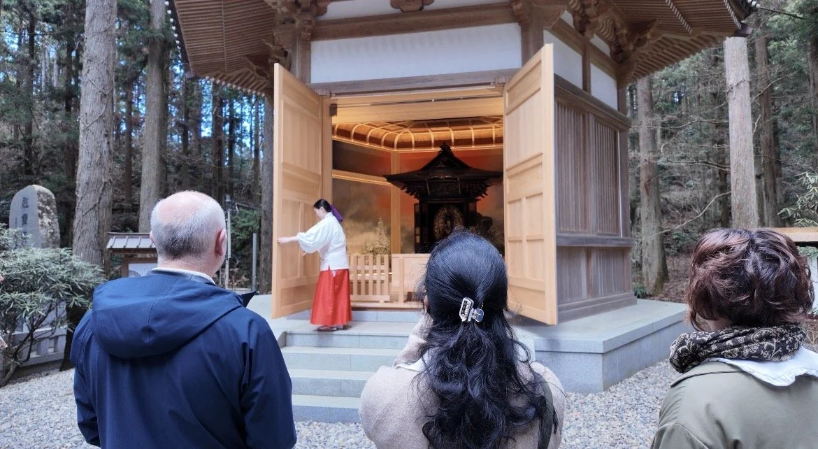 A small traditional Japanese shrine with a wooden exterior and open doors, located in a wooded area. Inside, a priest in white and red robes performs a ritual as three visitors watch.