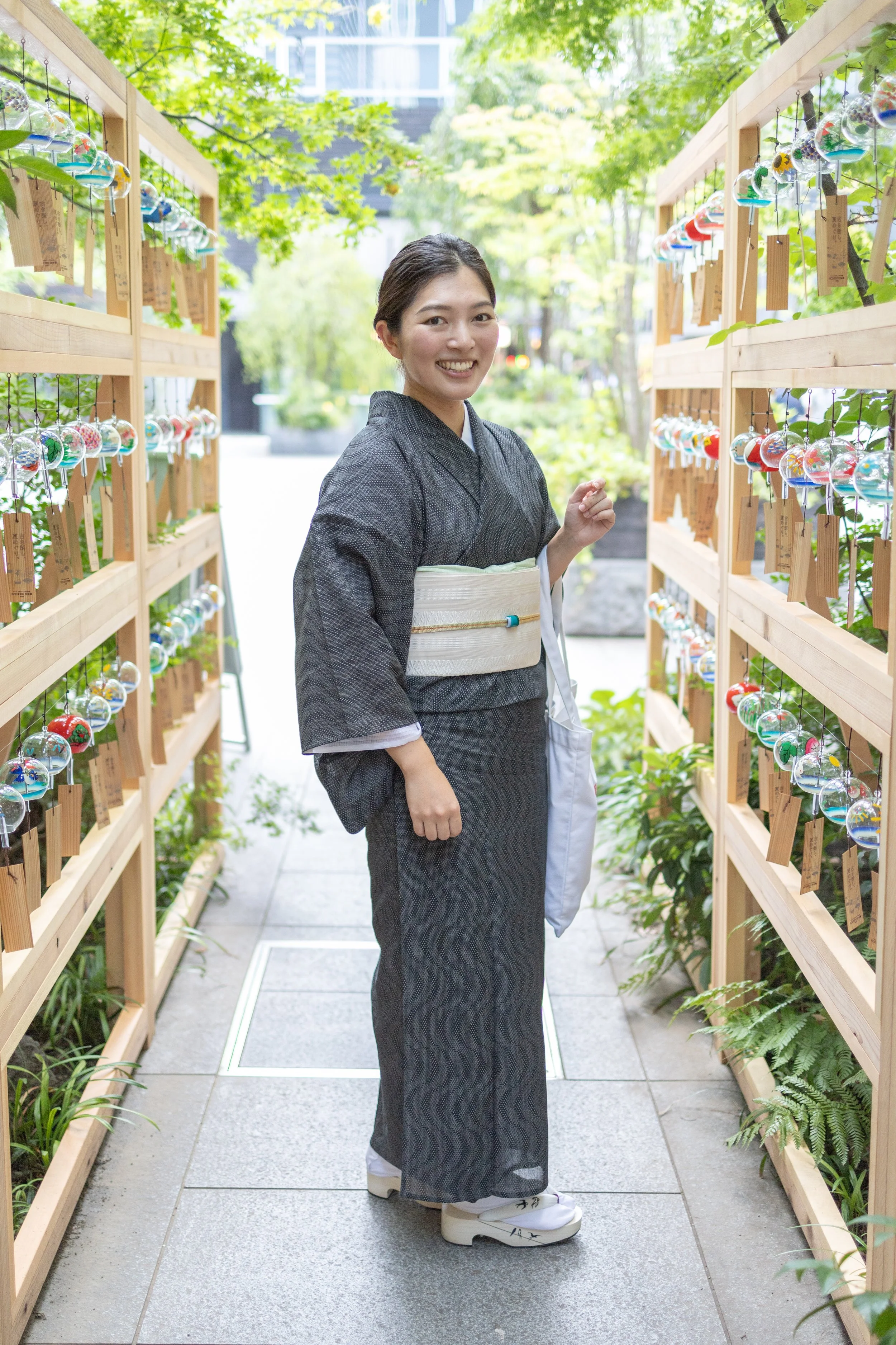 A smiling woman in traditional Japanese kimono standing between wooden display shelves with hanging glass wind chimes on a sunny outdoor pathway.