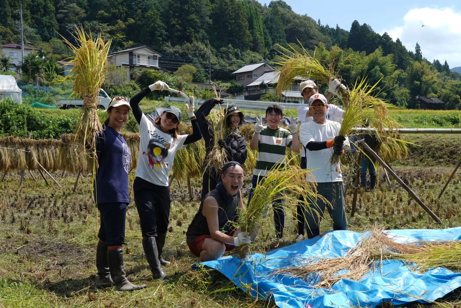 Group of people harvesting rice in a field, smiling and posing for the camera. They are holding bundles of harvested rice and are dressed in casual farm work clothes, with some wearing gloves and boots.