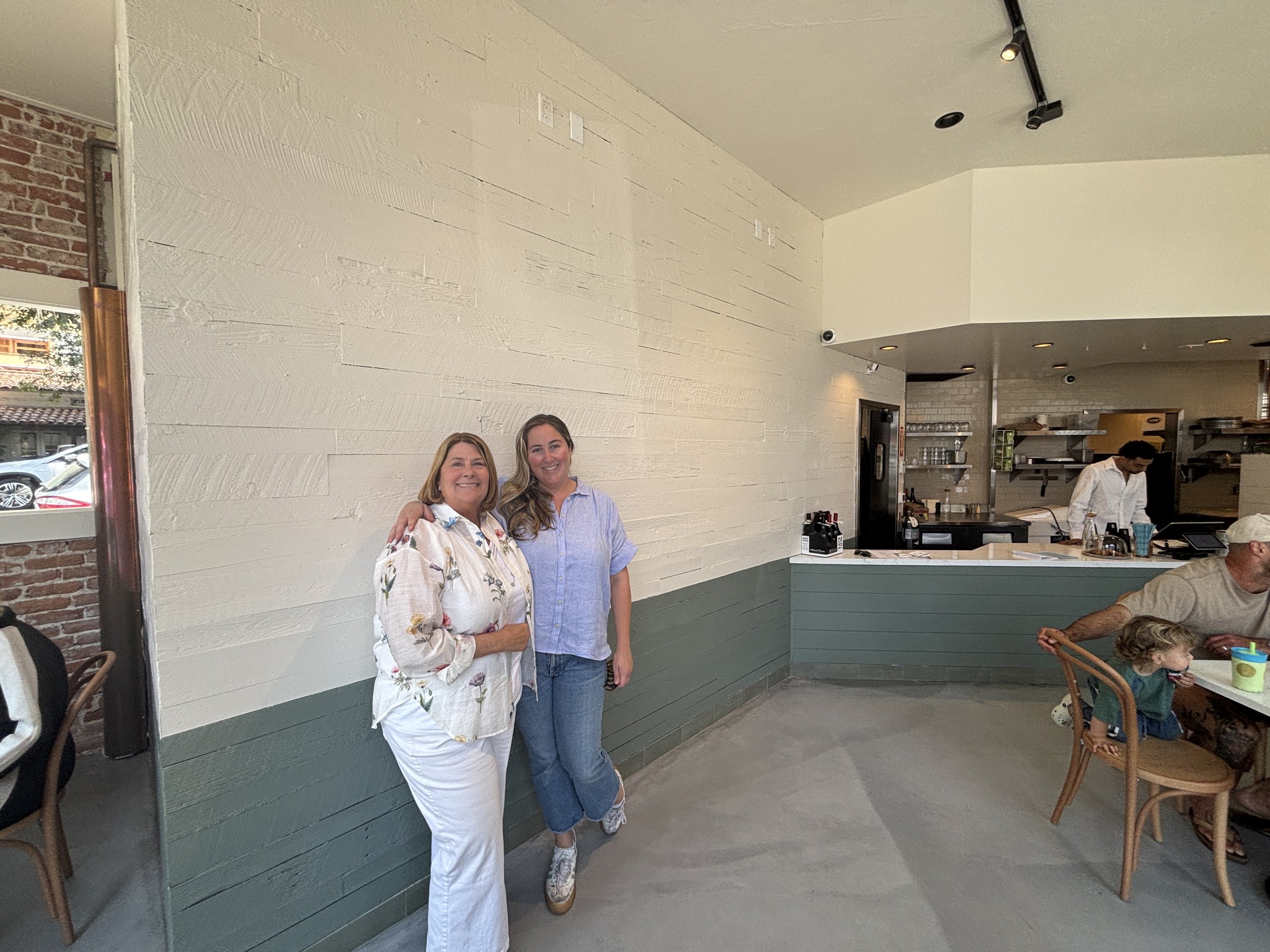 Two women standing together inside a restaurant, smiling at the camera. One wears a white floral shirt, and the other wears a light blue shirt. In the background, there is a kitchen area with shelves and a chef preparing food, and a child sitting at a table.