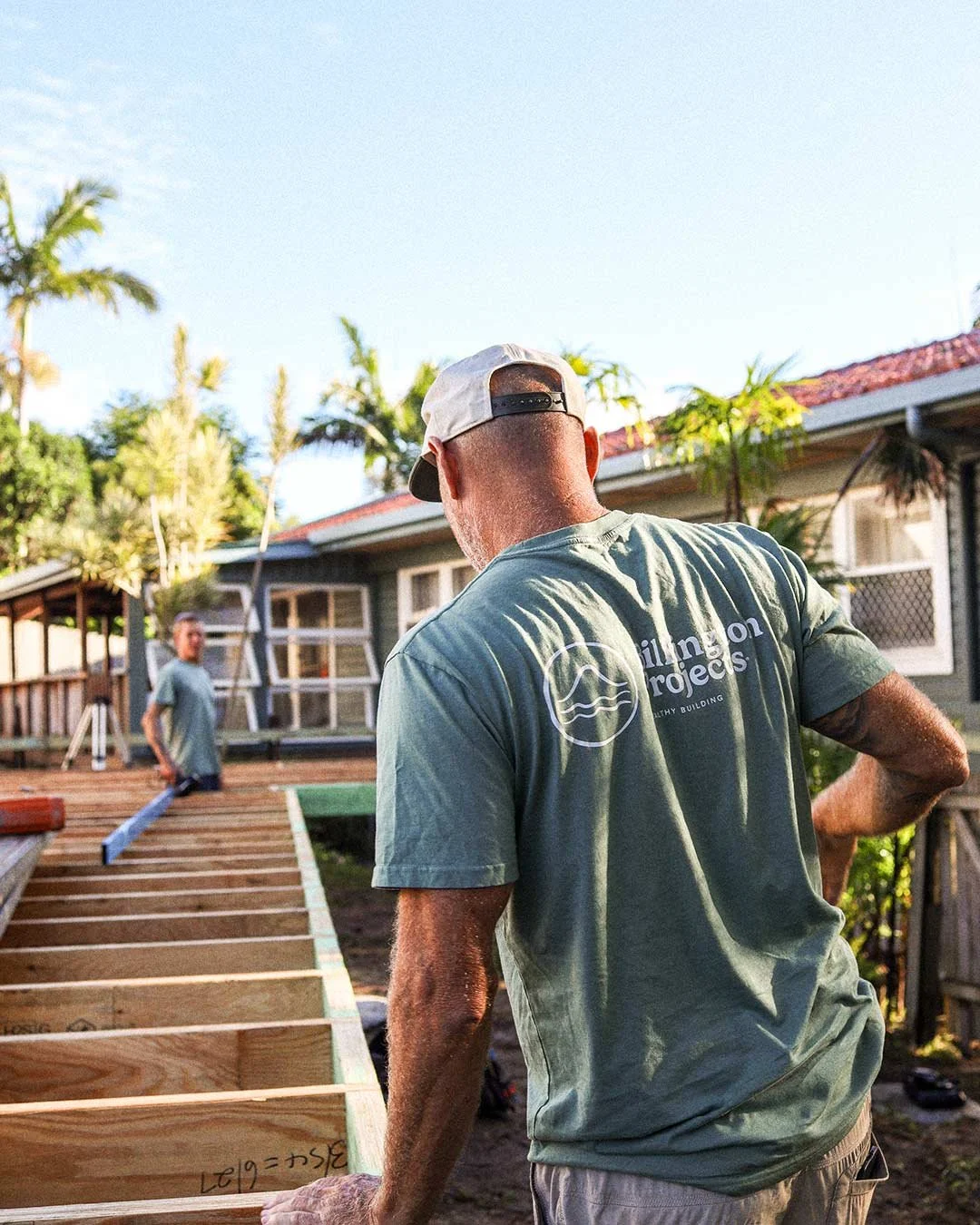 Two men working on a wooden deck construction outdoors, with one man in the foreground and the other in the background, surrounded by trees and a house.