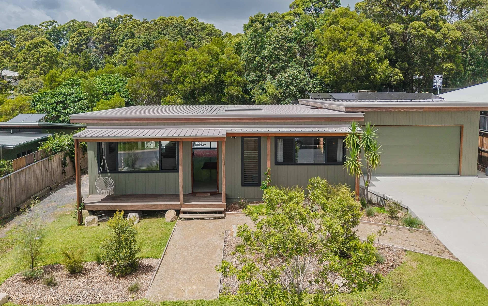 A modern single-story house with a potted plant, small porch, and a yard with young shrubs and trees, surrounded by a wooden fence, and with a background of lush greenery and trees.