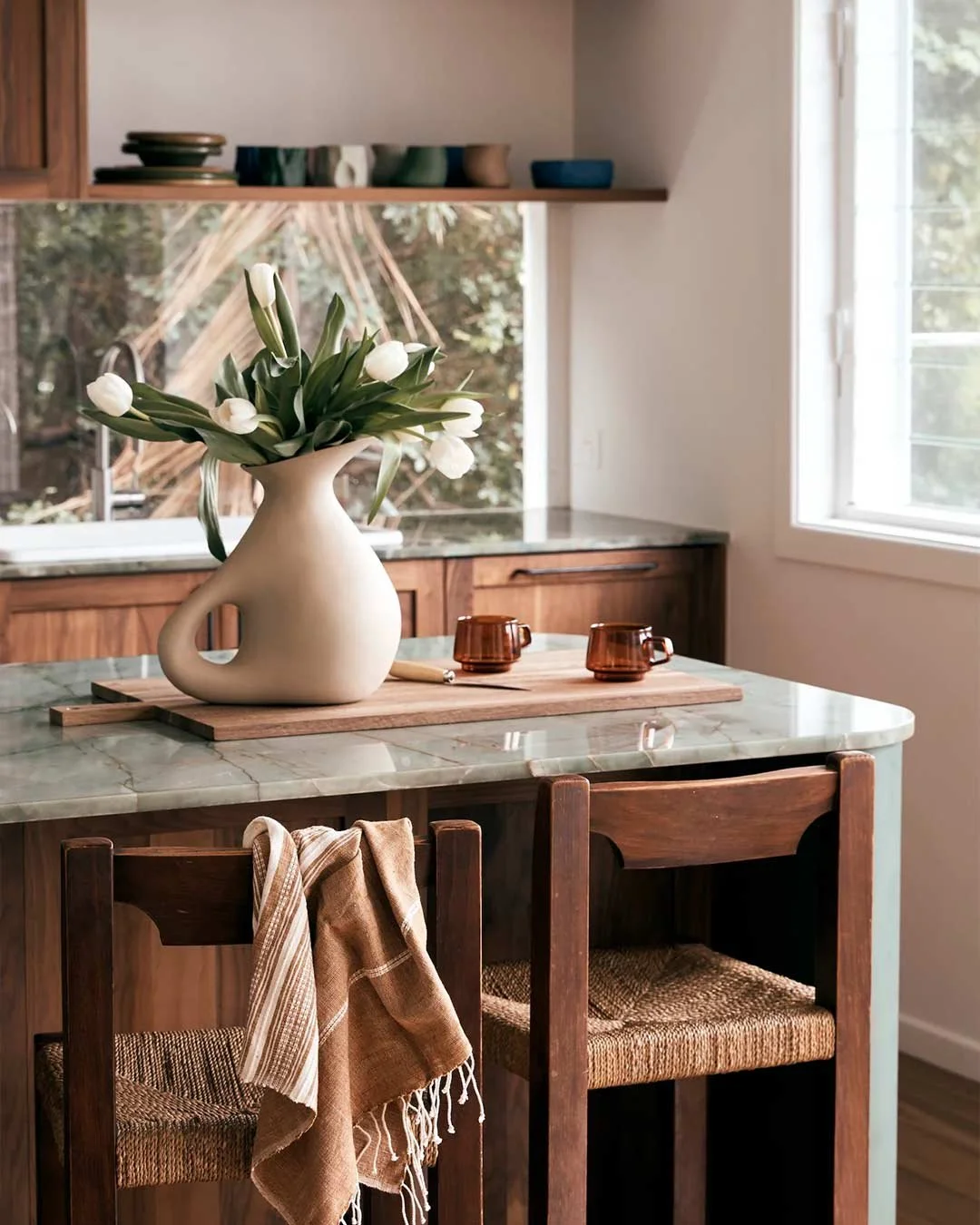 A kitchen with a marble countertop, wooden cabinets, and a window. On the counter, there is a beige vase with white tulips, two small amber cups, and a wooden tray. Two wooden chairs with woven seats are positioned at the counter, with one chair holding a folded beige and white striped cloth. Outside the window, trees and a wooden structure are visible.