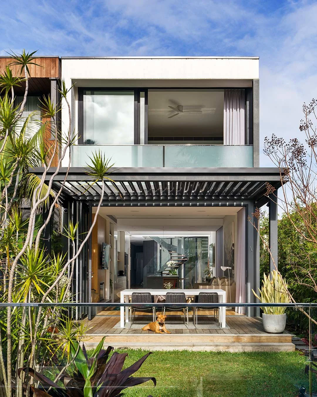 Modern multi-story house with large glass windows and a balcony, surrounded by greenery, with a dog lying on the porch.