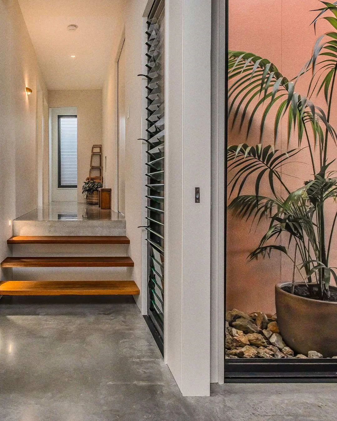 Interior of a modern house with a small staircase with wood steps, a sliding door, potted plants, and concrete flooring.