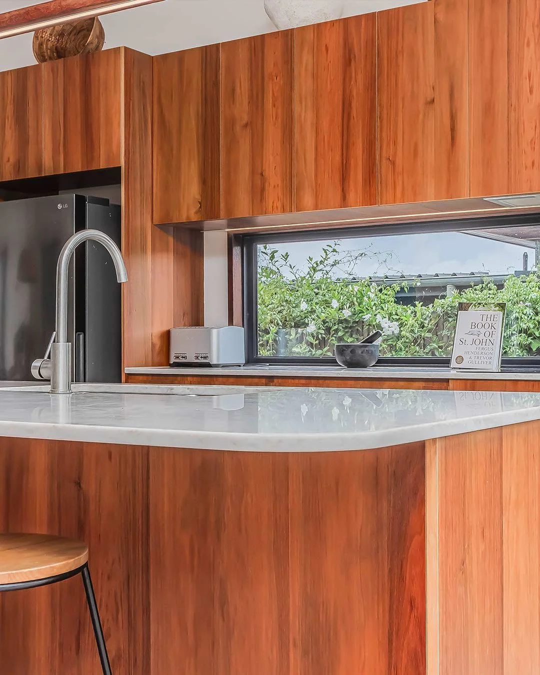 A kitchen counter with a marble countertop, a stainless steel sink with a tall faucet, a small toaster, a black bowl with a spoon, a framed book titled 'The Book of St. John,' and a window showing greenery outside.
