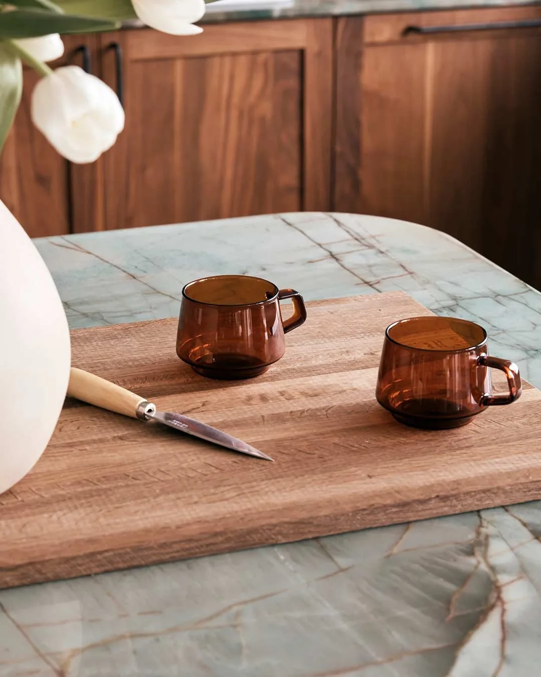 Two amber glass cups on a wooden cutting board, with a kitchen knife nearby, on a marble table with a wooden backsplash in the background.