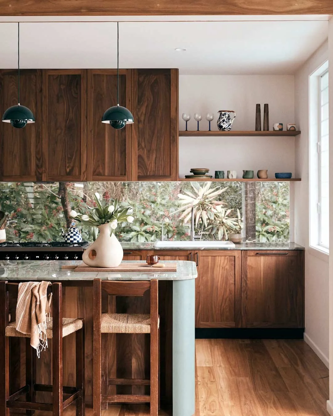 Modern kitchen with wooden cabinets, open shelves with dishes and vases, a vase with flowers on the marble island, and a view of greenery outside the window.