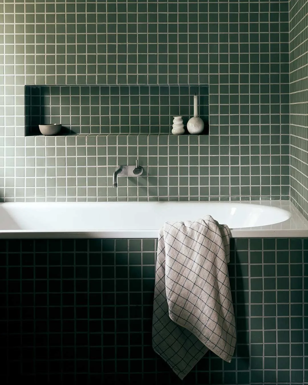 Green tiled bathroom with a built-in shelf holding minimal white decor, a white bathtub with a beige checkered towel hanging over it, and a wall-mounted faucet.