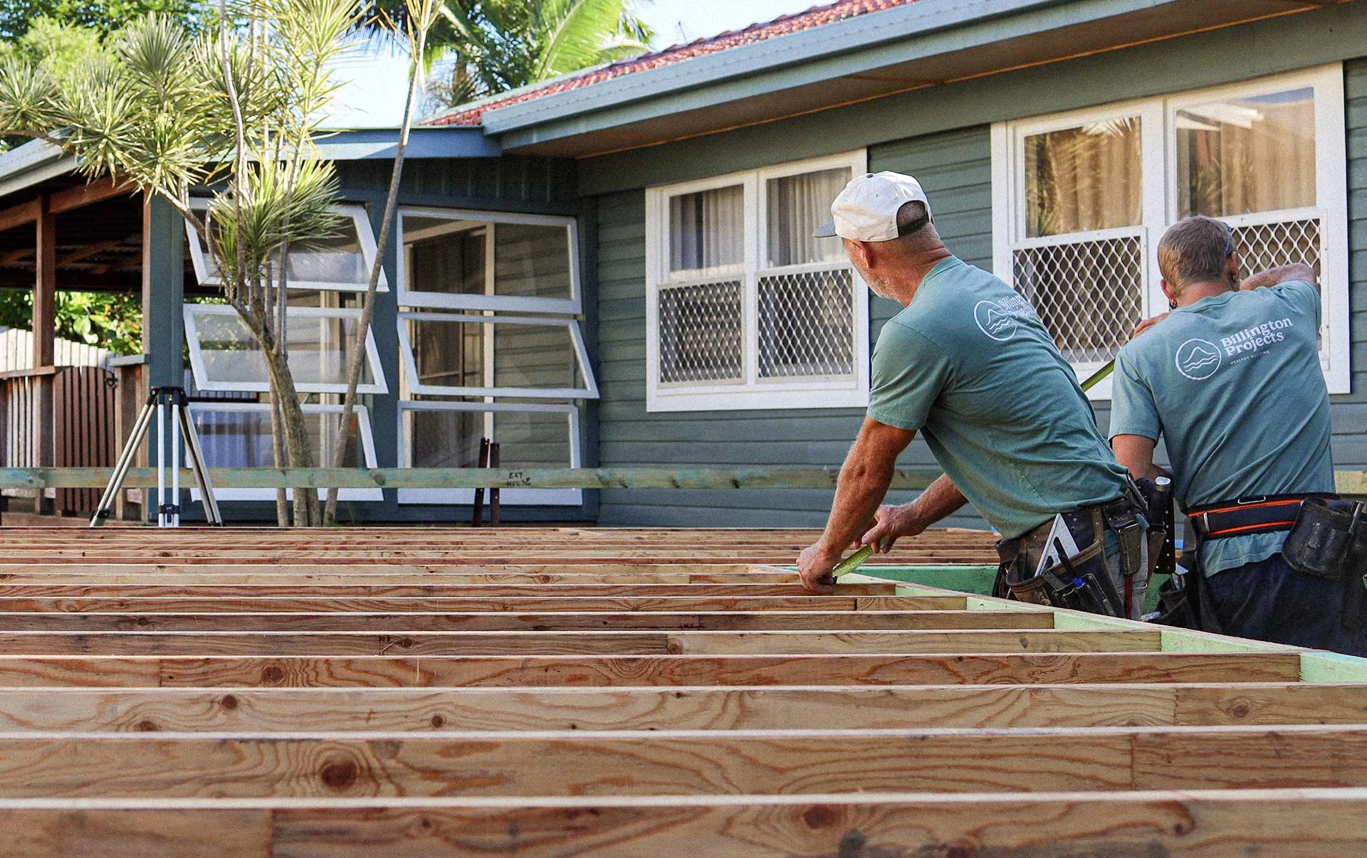 Two construction workers are working on a wooden deck in front of a green house with white framed windows. They are measuring the deck's dimensions, and the scene includes a tree and some construction equipment.