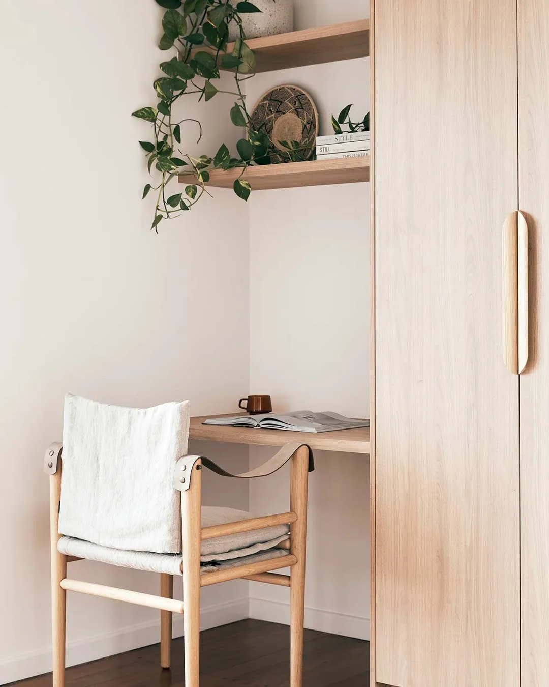 A minimalist workspace with a wooden desk, a white chair with a fabric seat, and a built-in wooden cabinet. There are two floating shelves above, decorated with a potted plant, a decorative plate, and a few books.