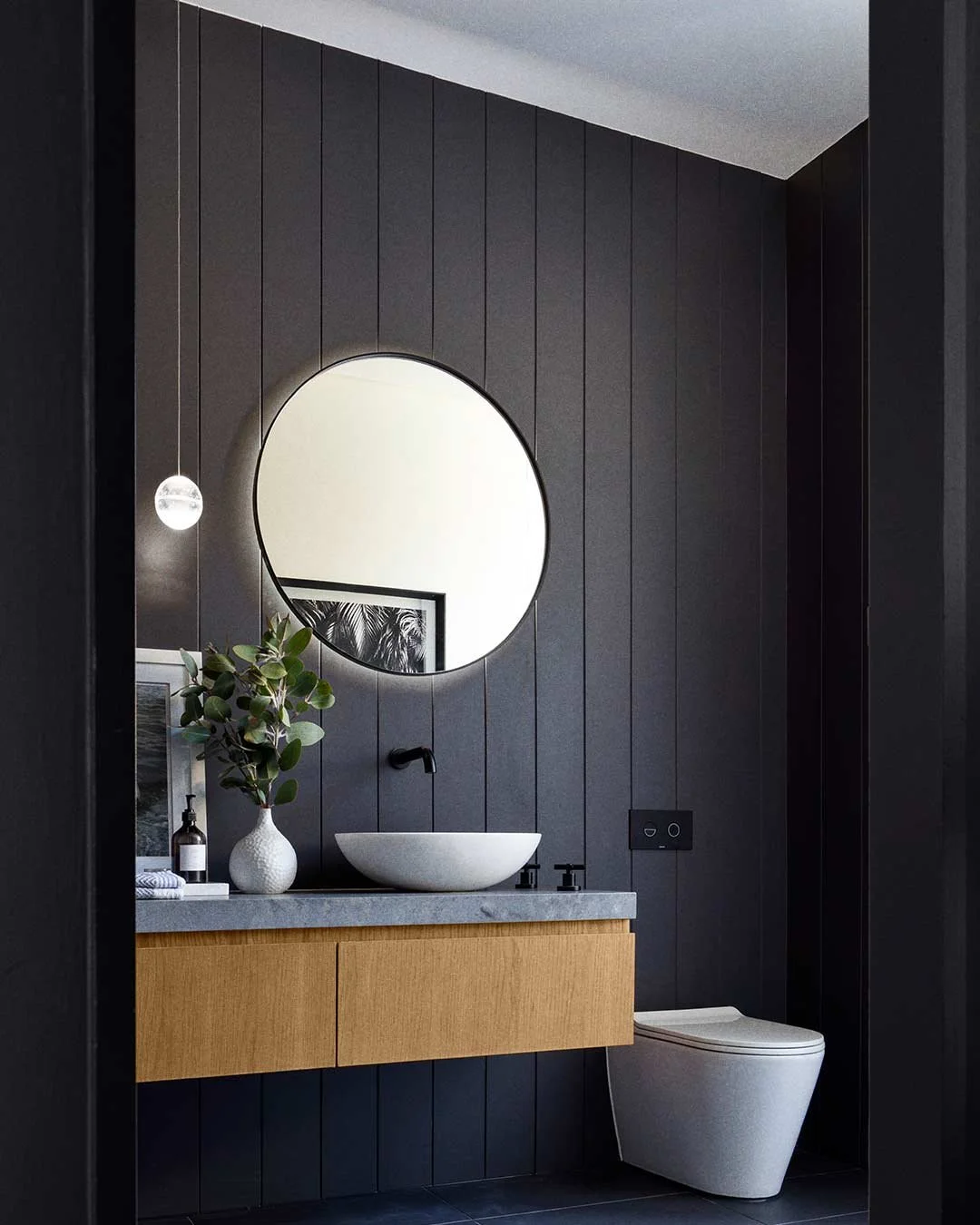 Modern bathroom with black wooden paneled wall, round mirror, white vessel sink on a gray stone countertop, wooden vanity, decorative plant, and white toilet.