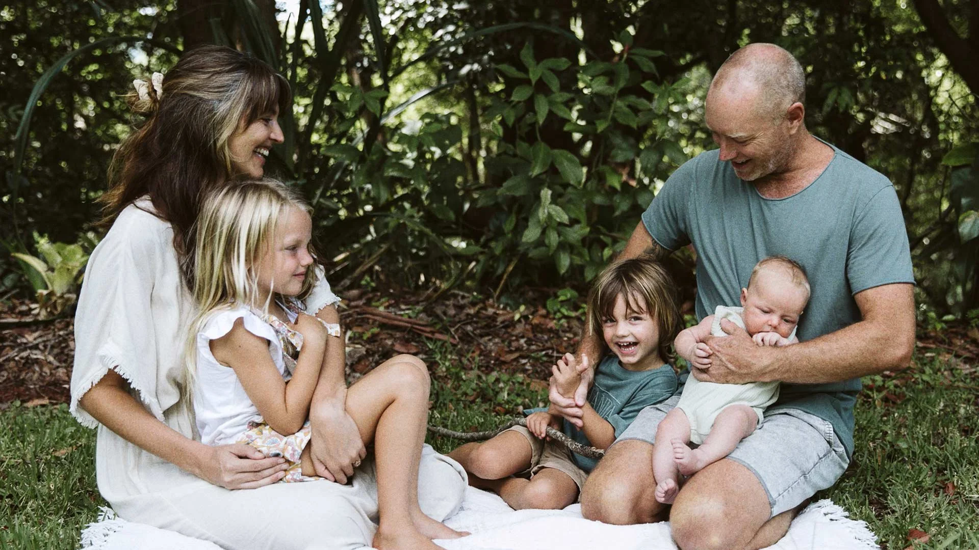 A family of six enjoying time together outdoors in a forest setting, with a woman, a man, and four children sitting on a blanket, smiling and laughing.