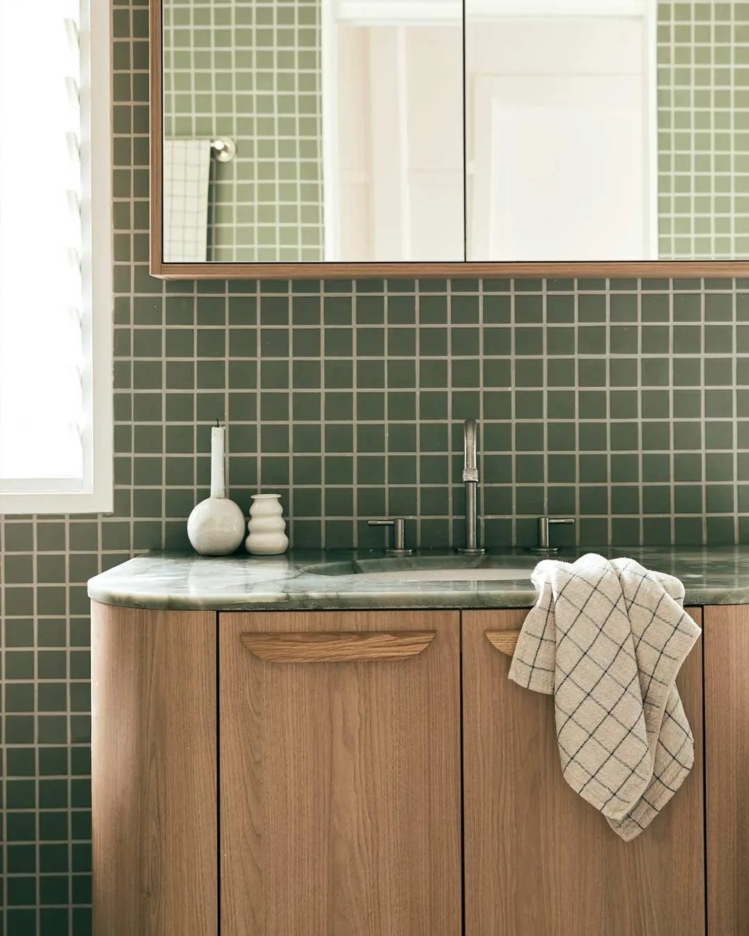 Bathroom vanity with green tiled wall, marble countertop, wooden cabinet, mirror, white best, and checkered towel.