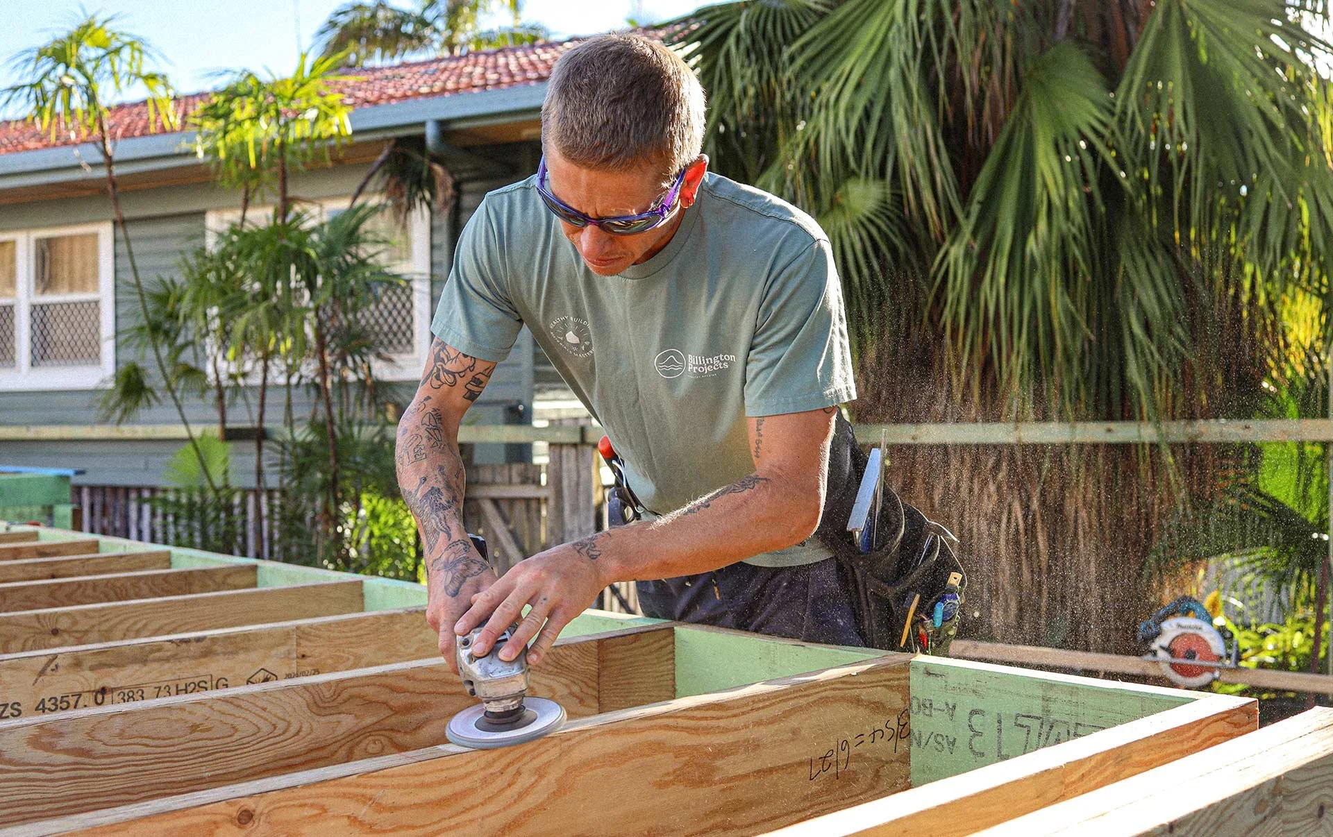 A man with tattoos on his arms and wearing sunglasses is sanding a wooden structure outdoors during daytime. There are trees, a house with a red roof, and a cloudy sky in the background.