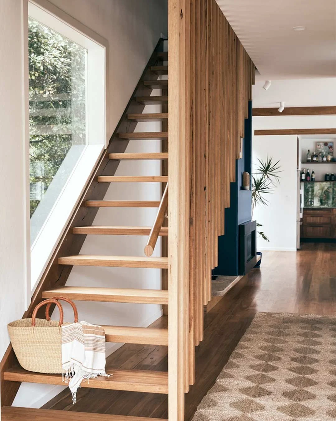 Wooden staircase with open risers next to a large slanted window, inside a modern home with wooden accents and a patterned rug on the floor.