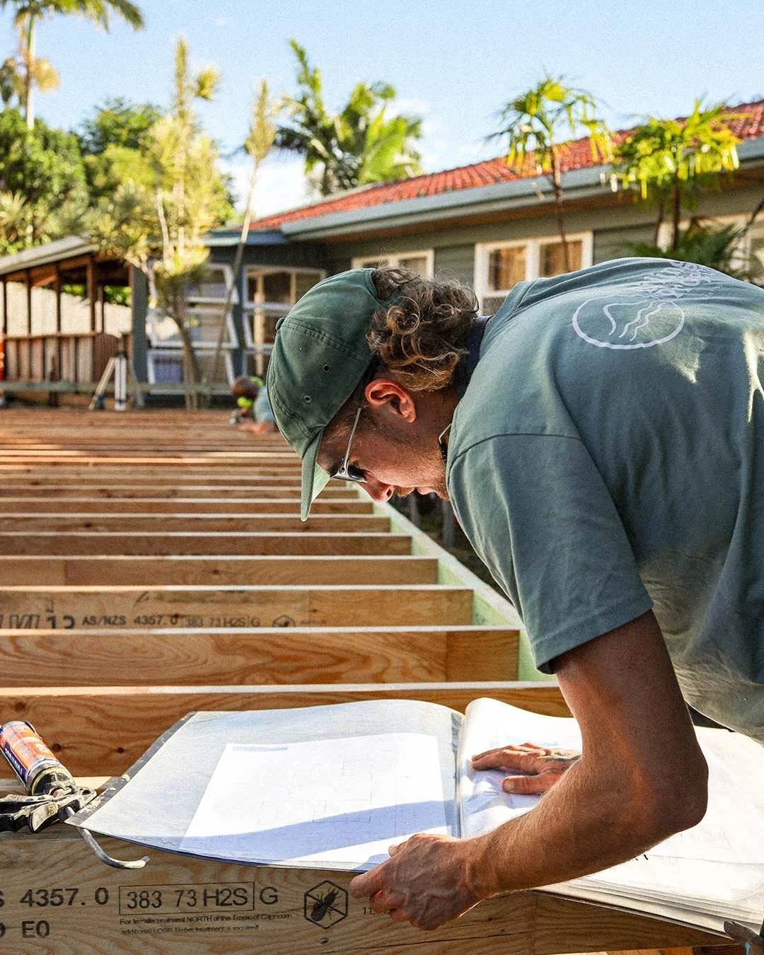 A man in a green cap and glasses working on a wooden deck foundation, reviewing blueprints on a table outside a house, with some plants and trees in the background.