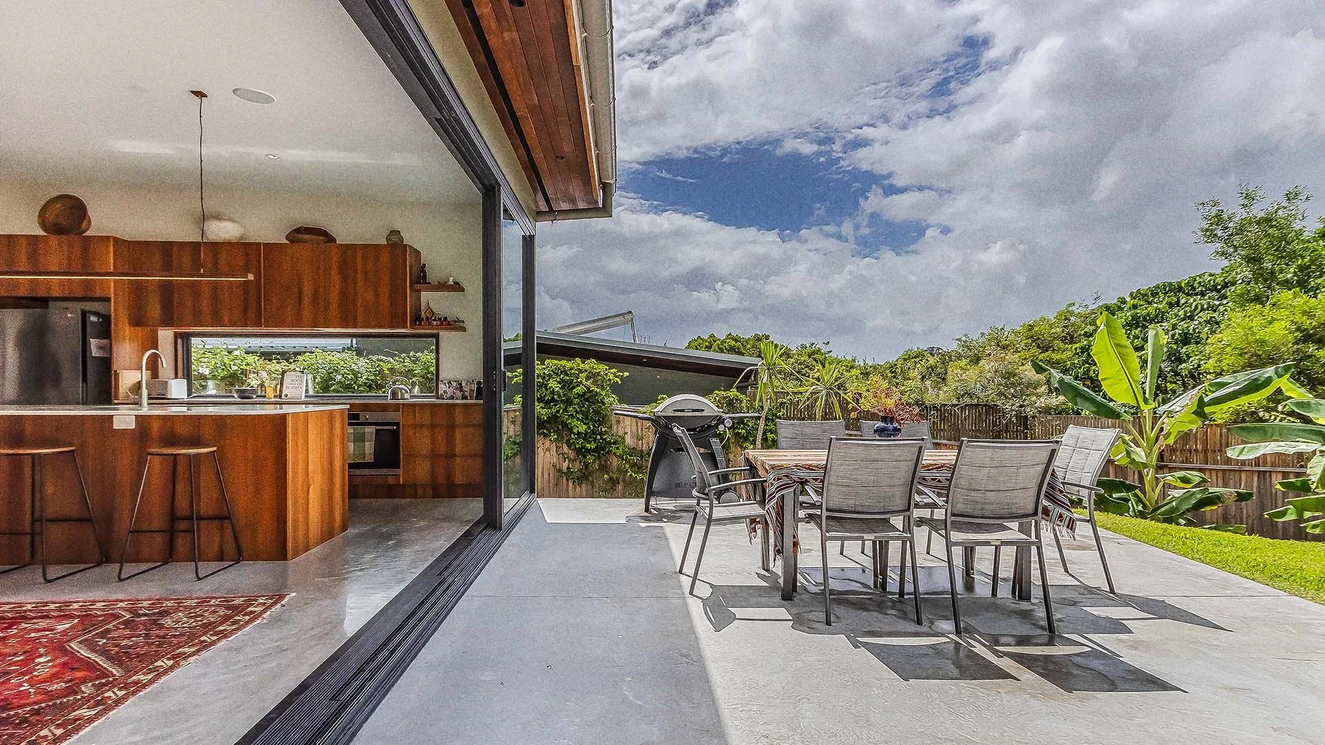Open living room with a view to a patio, featuring a dining table and chairs, a barbecue grill, and outdoor greenery under a cloudy sky.