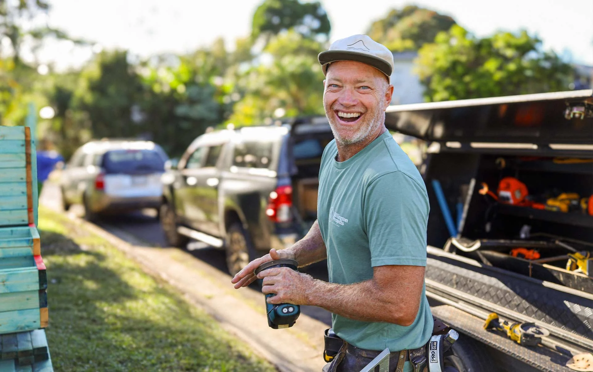 A smiling man in work clothes holding a power tool outdoors in front of parked cars and a tool trailer, with trees in the background.