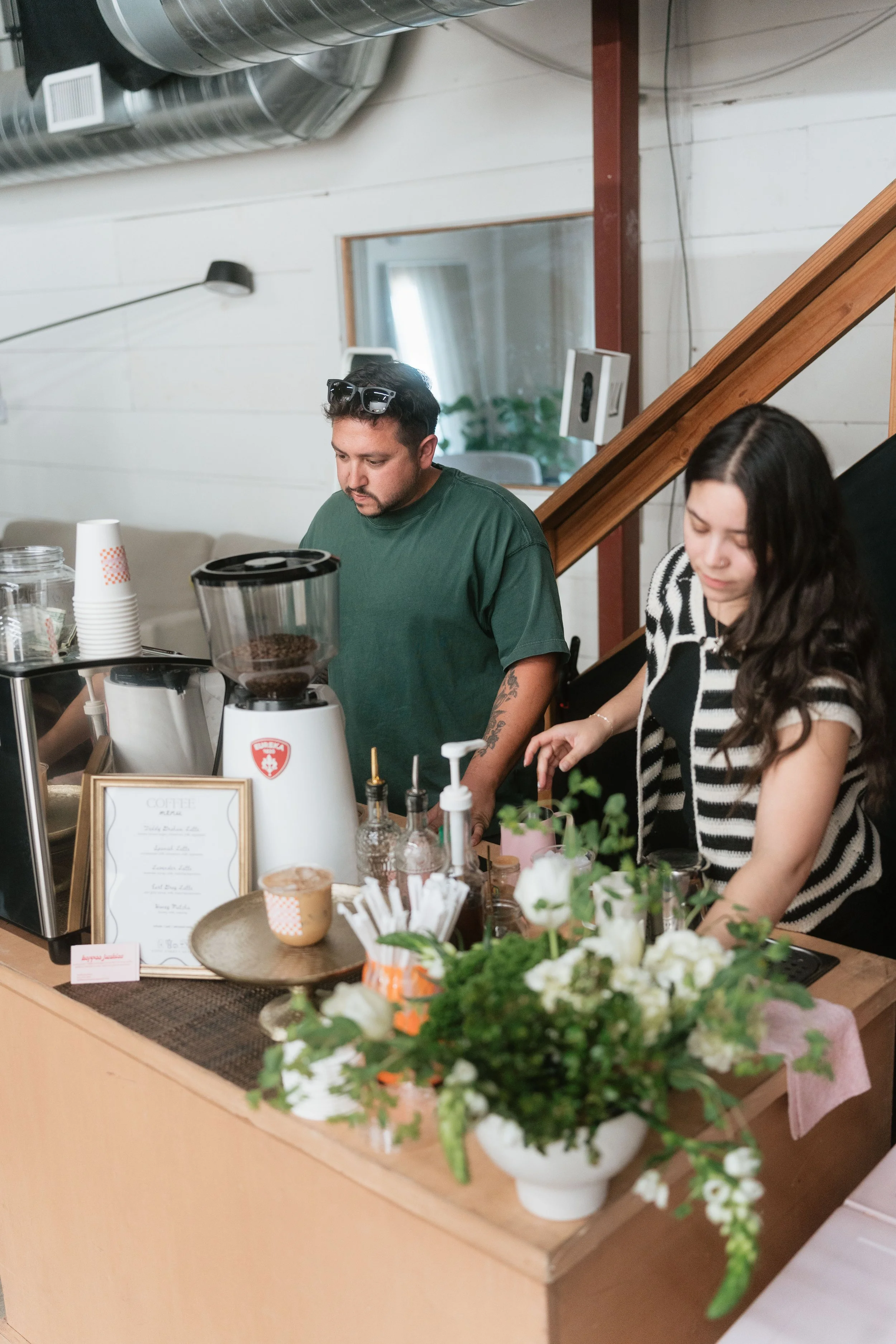People preparing coffee at a counter in a cafe with white walls, ductwork, and plants in the foreground.