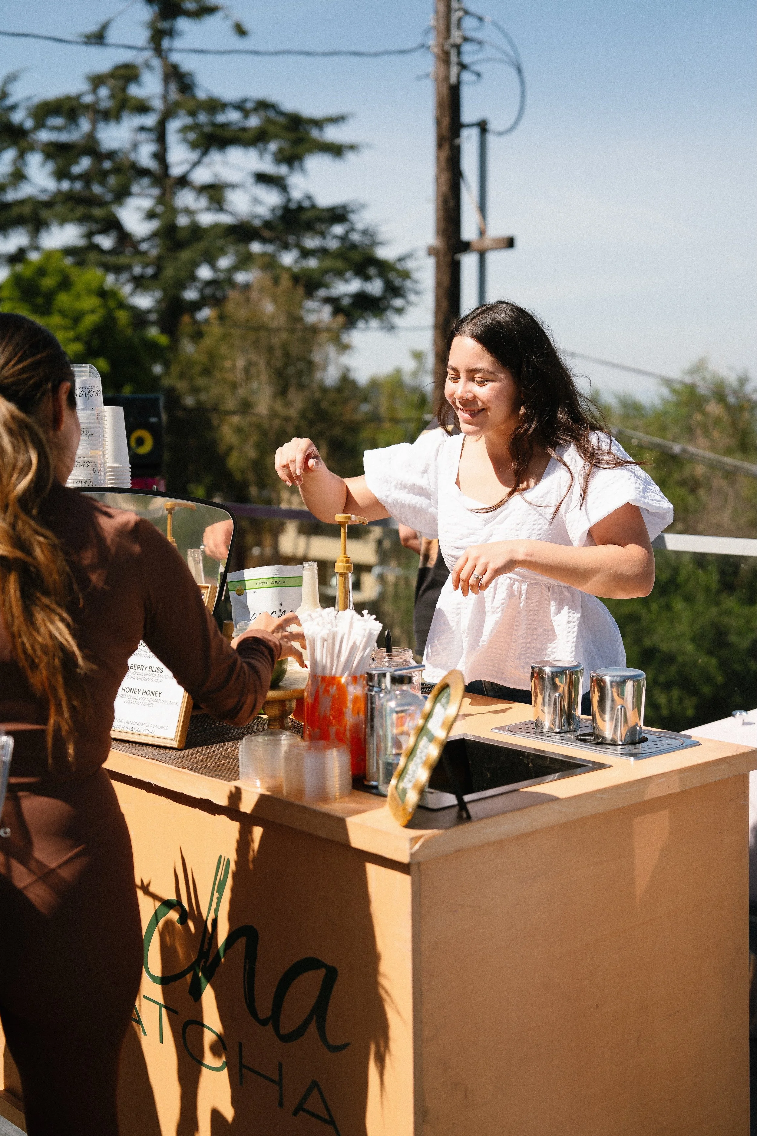 A woman with long dark hair wearing a white dress serving drinks at an outdoor booth with a sign that reads 'Cha'.