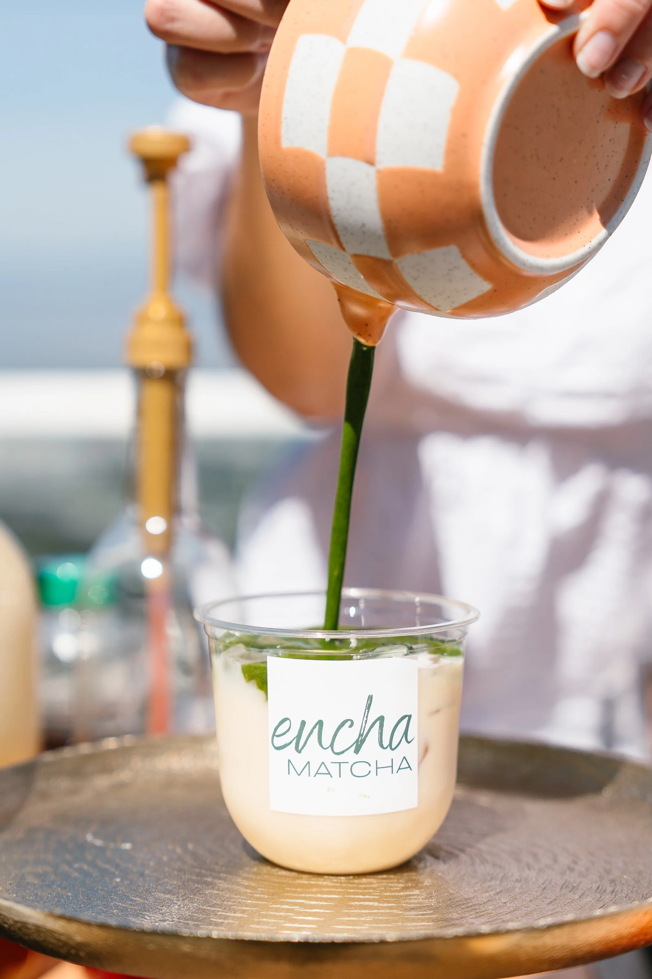 A hand pouring matcha latte with foam into a clear plastic cup with a sticker that says resca Matcha. The cup contains foam and matcha, and there are blurred objects in the background.