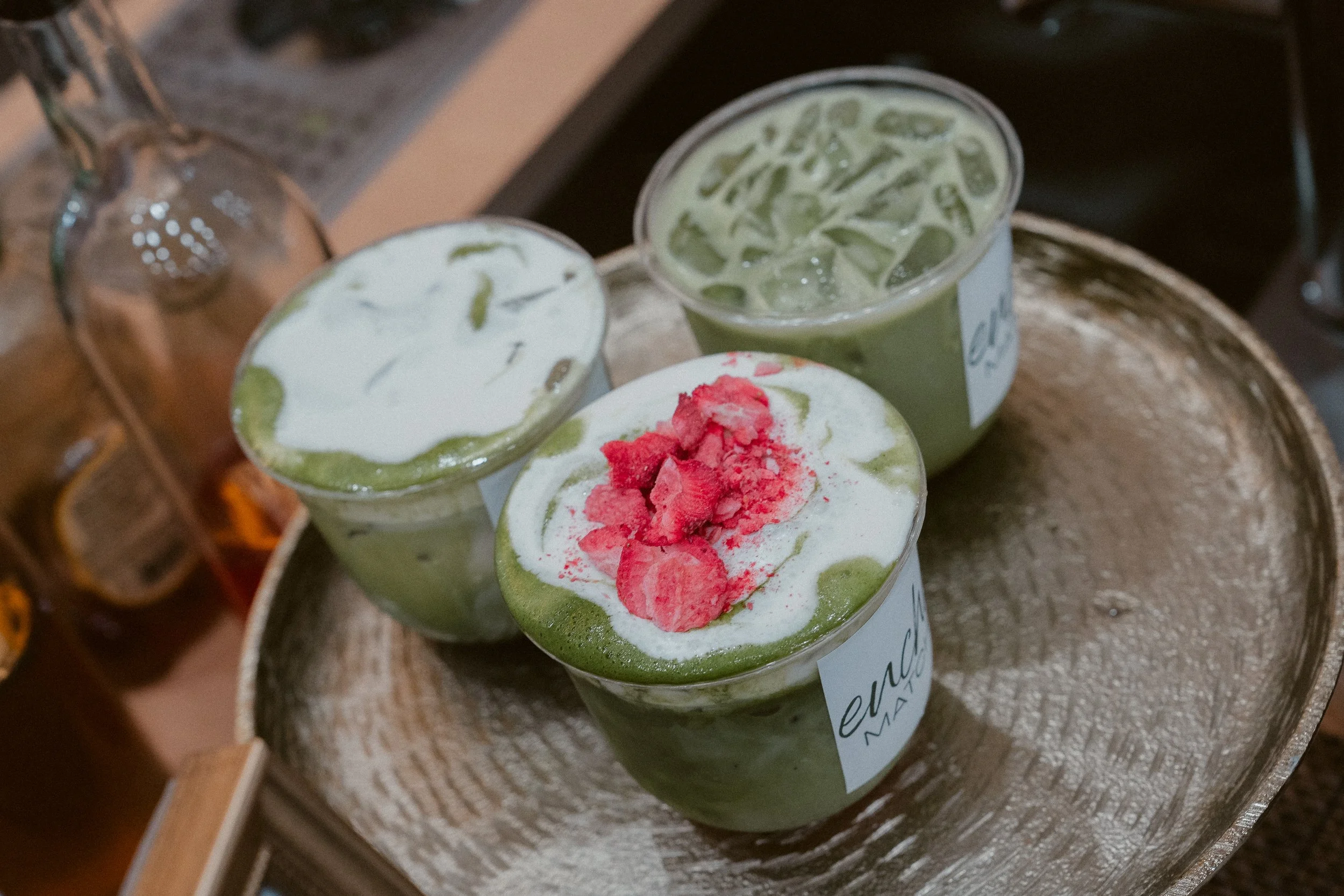 Three cups of matcha latte with whipped cream, one topped with pink crushed candy, on a wooden tray with a bottle in the background.