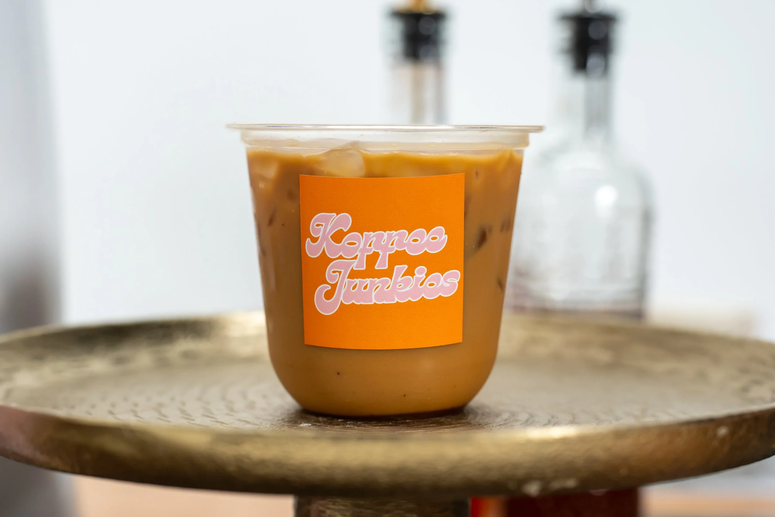 A plastic cup filled with iced coffee or tea and ice cubes, placed on a round wooden tray. The cup has an orange label with pink and white text that reads 'Kona Jumbies.'