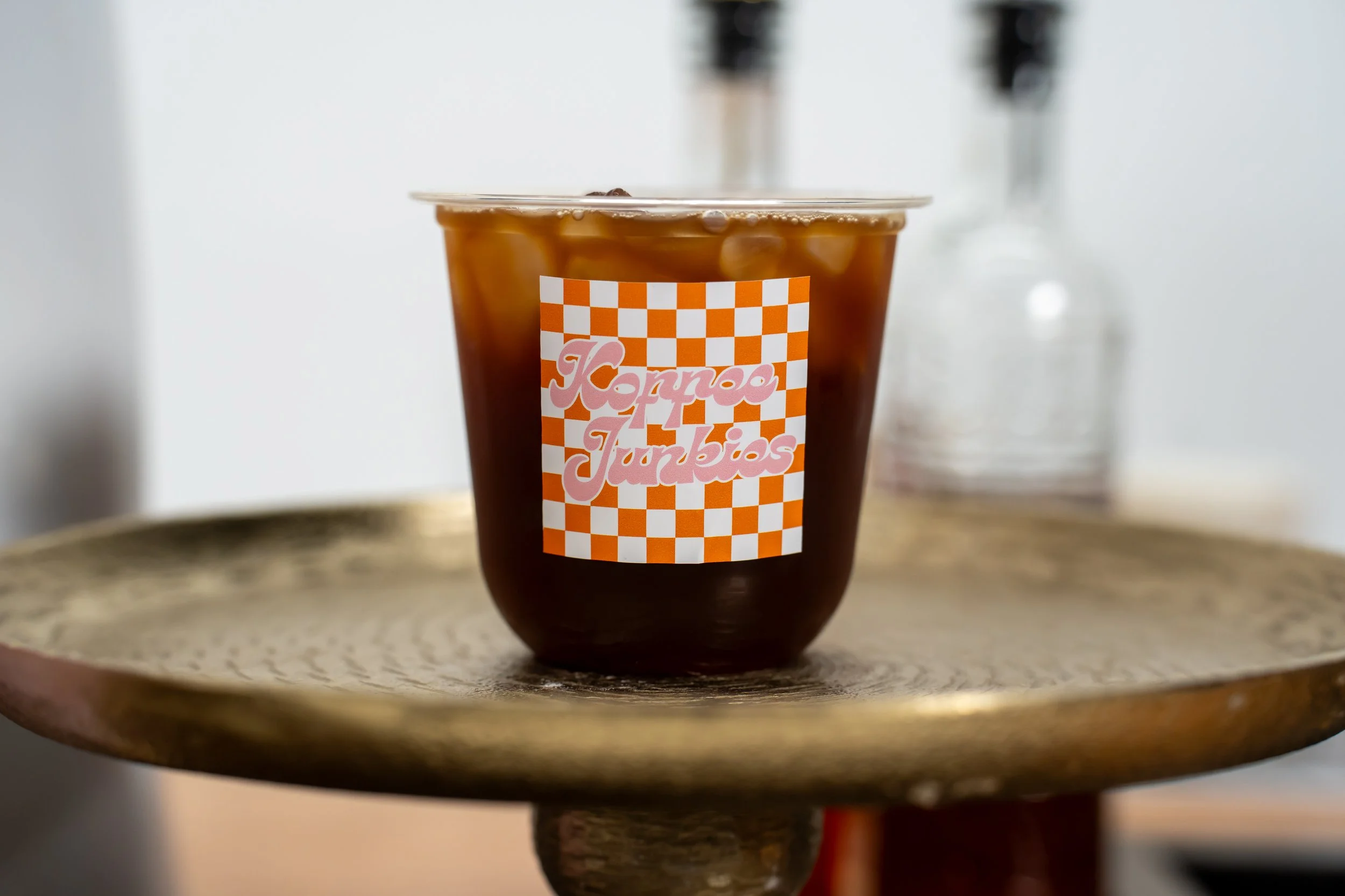 A brown iced drink in a clear cup with a checkered orange and white label that says 'Kono's Junkios,' sitting on a round, wooden tray.