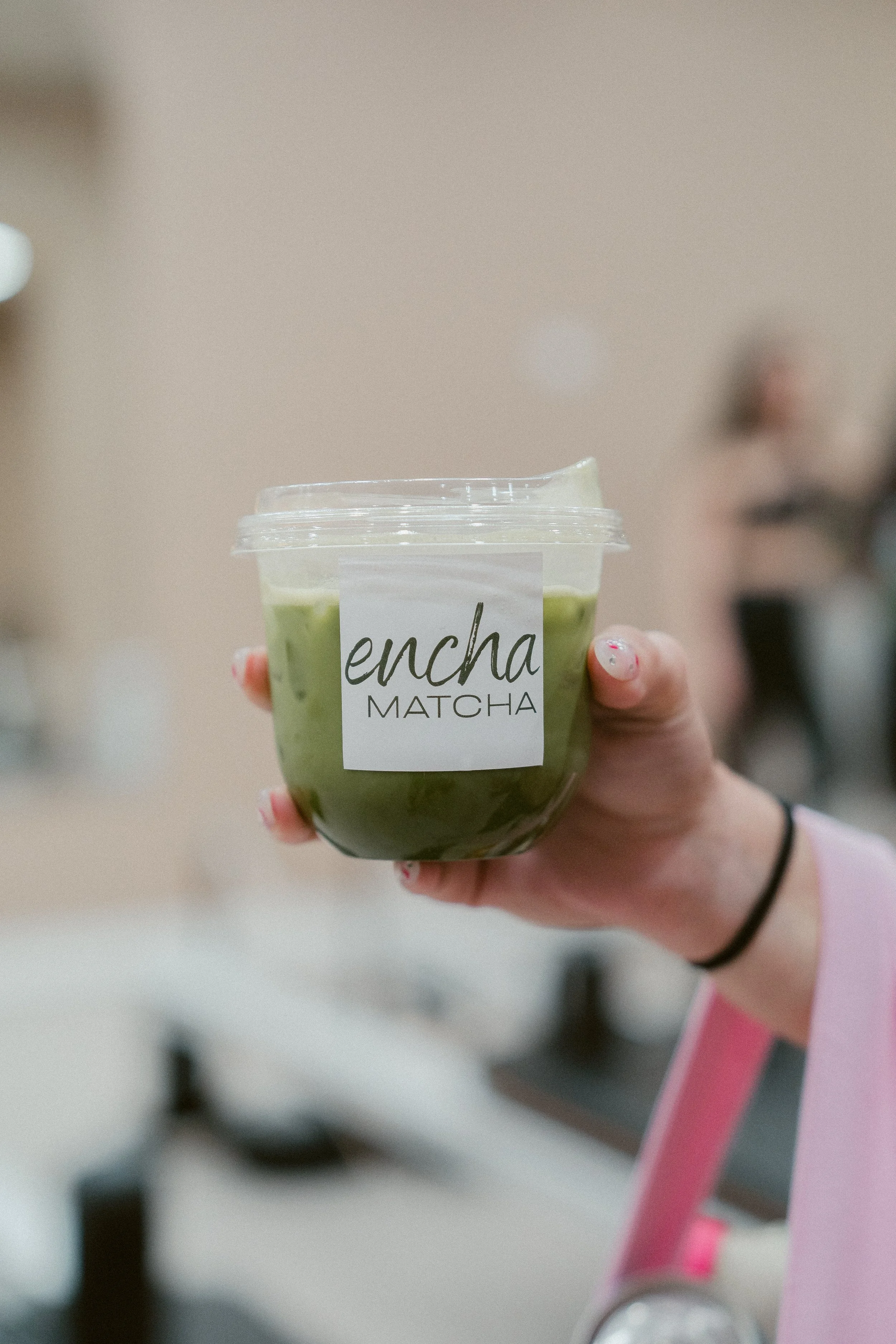 A hand holding a cup of matcha latte with a plastic lid, labeled 'Encha Matcha'.