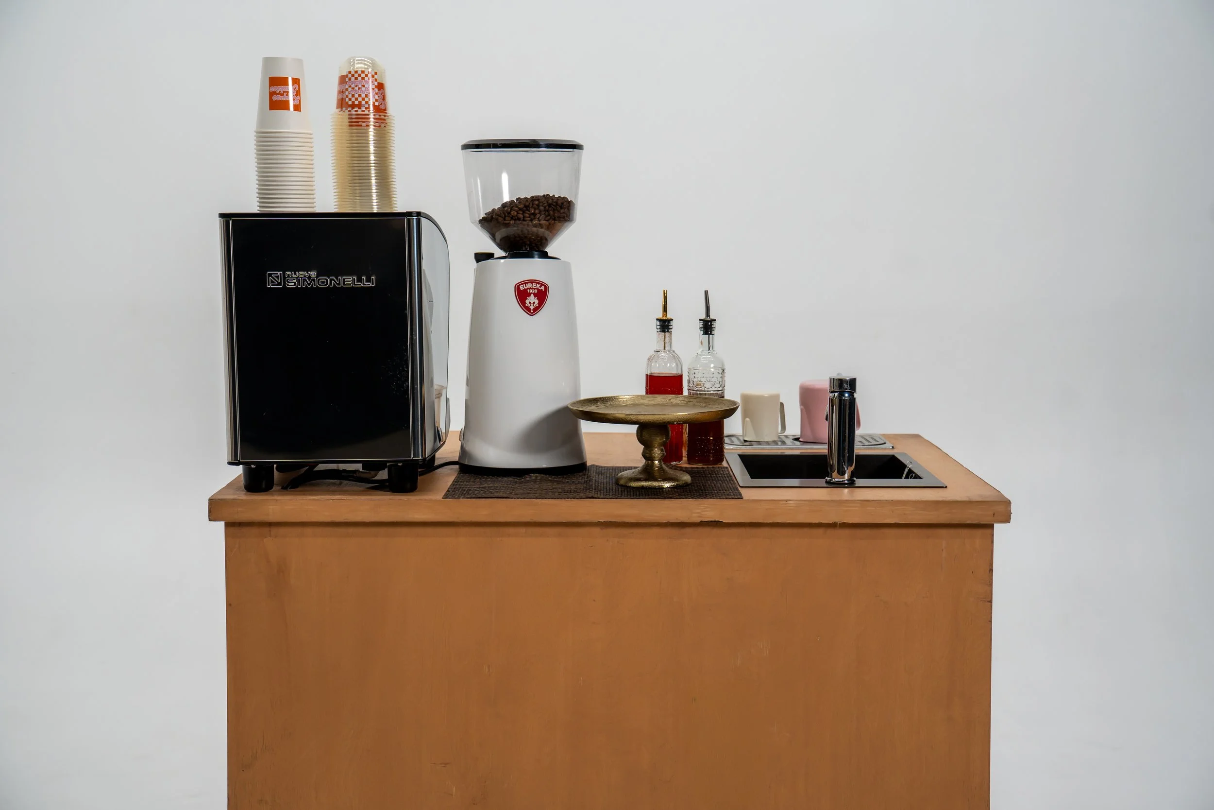 A wooden countertop with a coffee machine, a clear coffee bean grinder, several stacks of disposable cups, and various condiment bottles including syrup and cream.