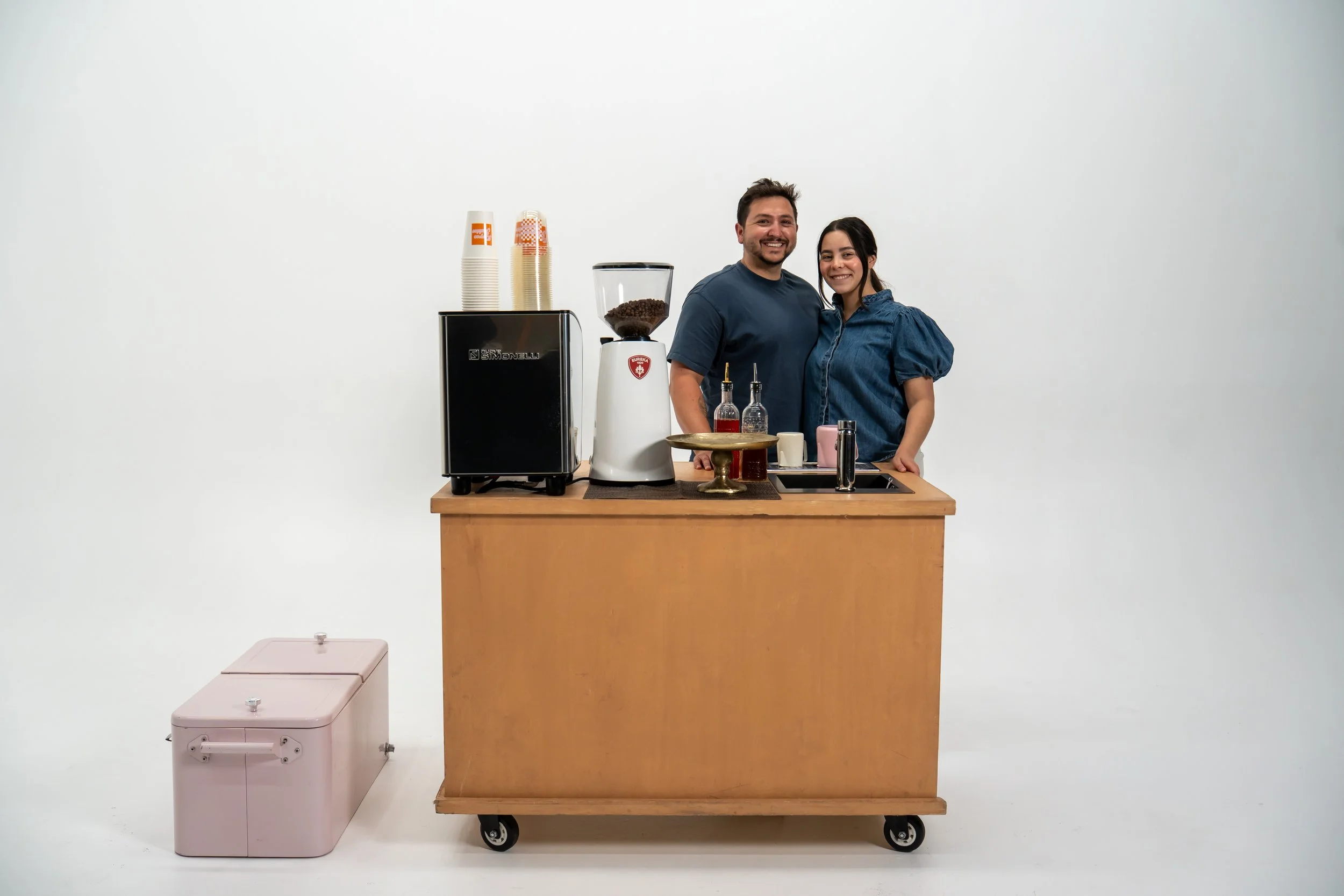 Two smiling baristas behind a small wooden bar on wheels, with a coffee grinder, syrup bottles, a pink cooler, and paper cups, against a plain white wall.