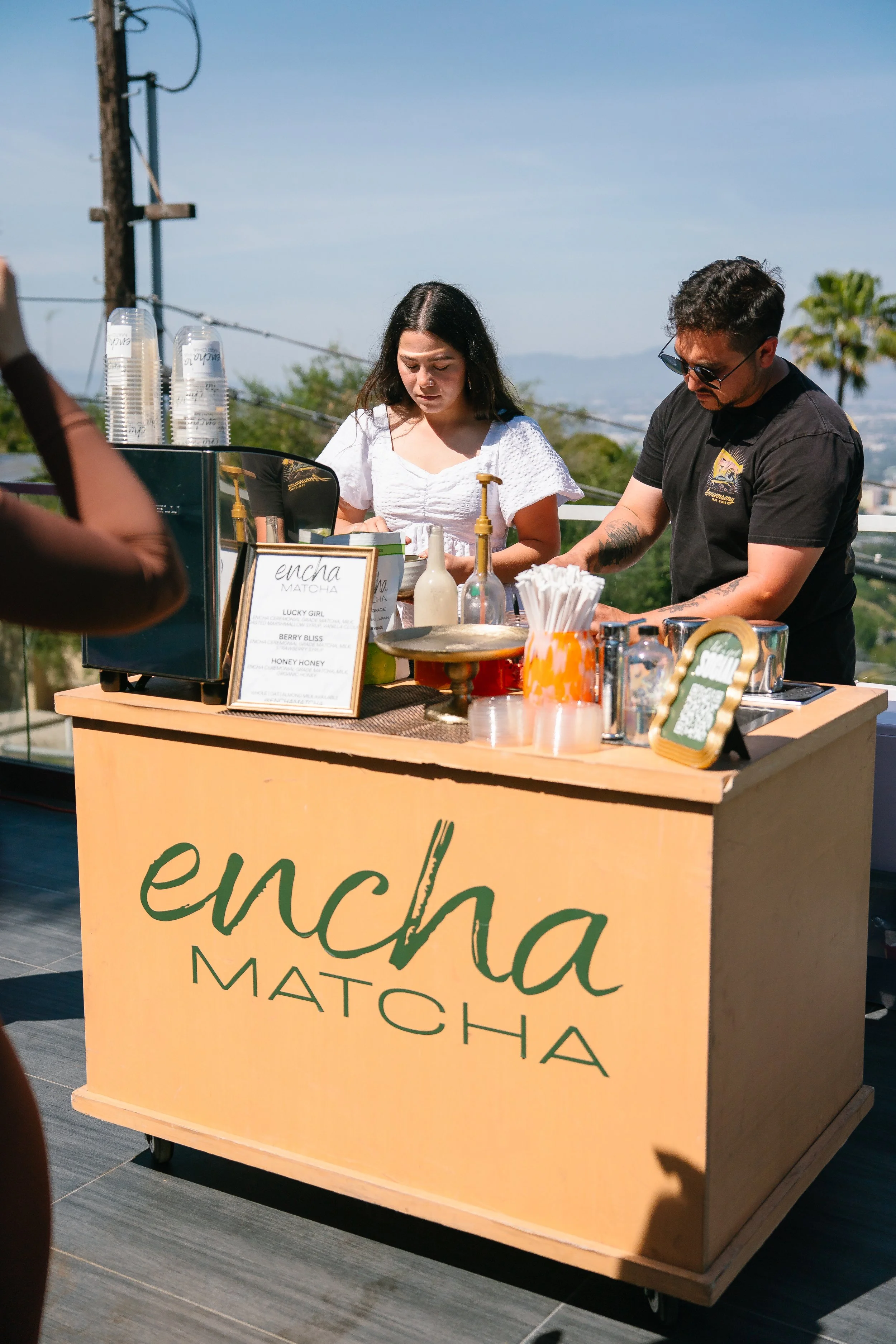 A food cart with a sign displaying the name 'Encha Matcha' and a menu listing matcha tea flavors. The cart is outdoors under a clear, sunny sky with trees and mountains in the background. Two people are working behind the cart, preparing drinks.