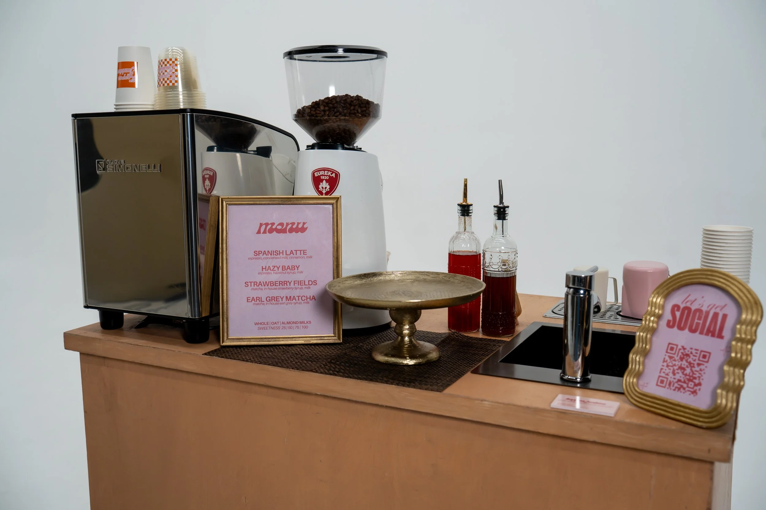 A countertop coffee station with a coffee grinder, jars of syrup, cups, and a framed menu listing various coffee drinks.