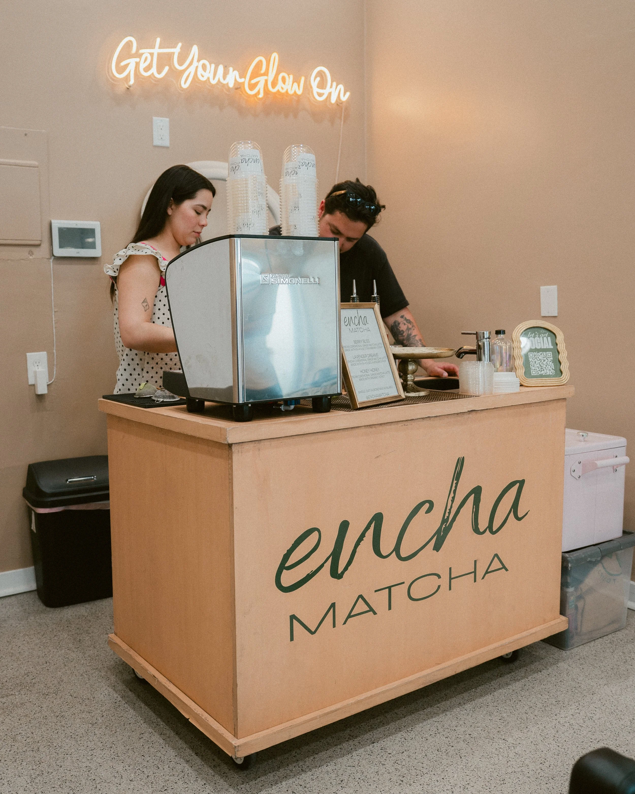 A wooden snack bar with a neon sign that says "Get Your Glow On," two servers behind the counter, a menu on a stand, condiment jars, and disposable cups.