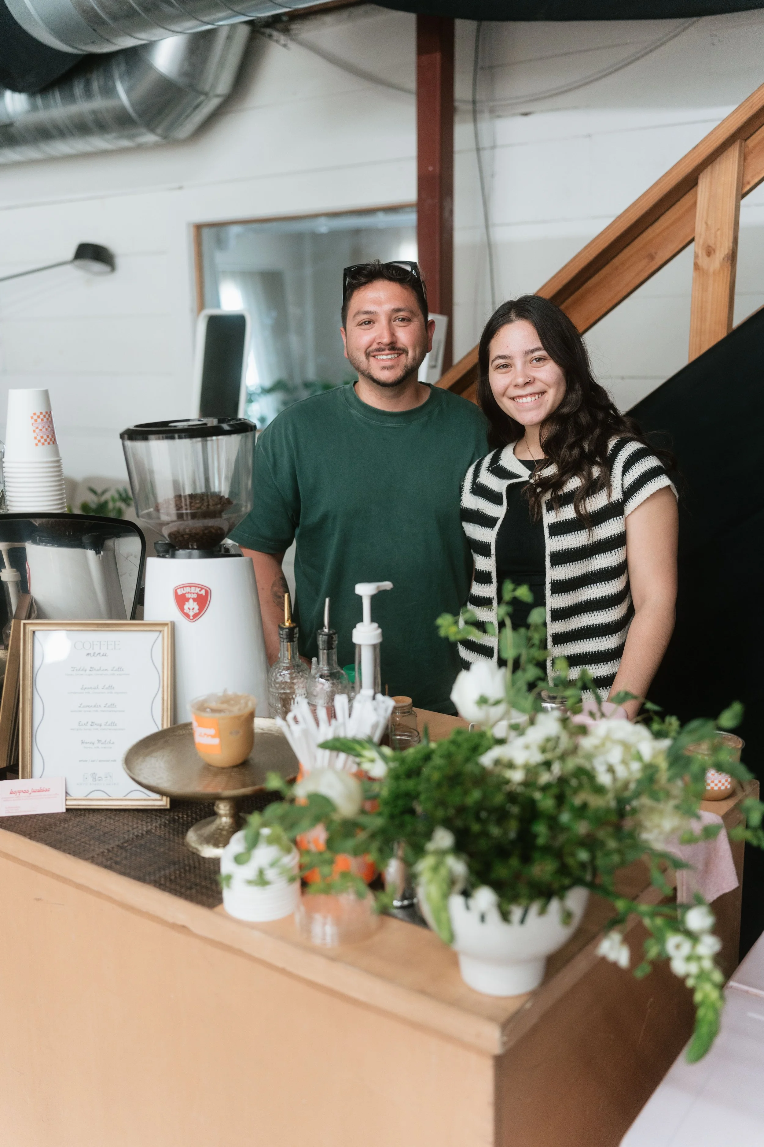 Two people smiling behind a coffee counter with coffee equipment, menu, and floral decorations in the foreground.