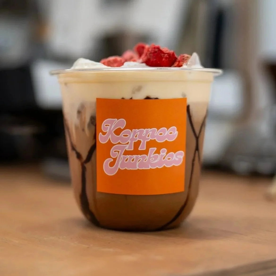 A cup of dessert topped with raspberries, chocolaty bits, and whipped cream, labeled 'Karpas Tumbias' on an orange sticker.