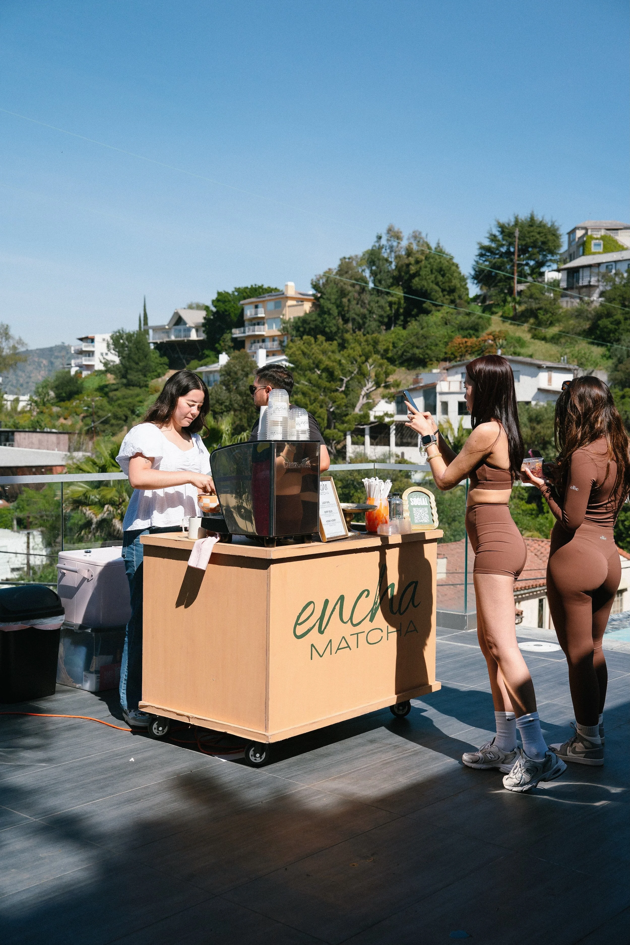Three women and one man are at an outdoor matcha beverage stand with a city hillside background. One woman is smiling at a laptop, the others are waiting, one is taking a photo, and the stand has cups and drinks on it.