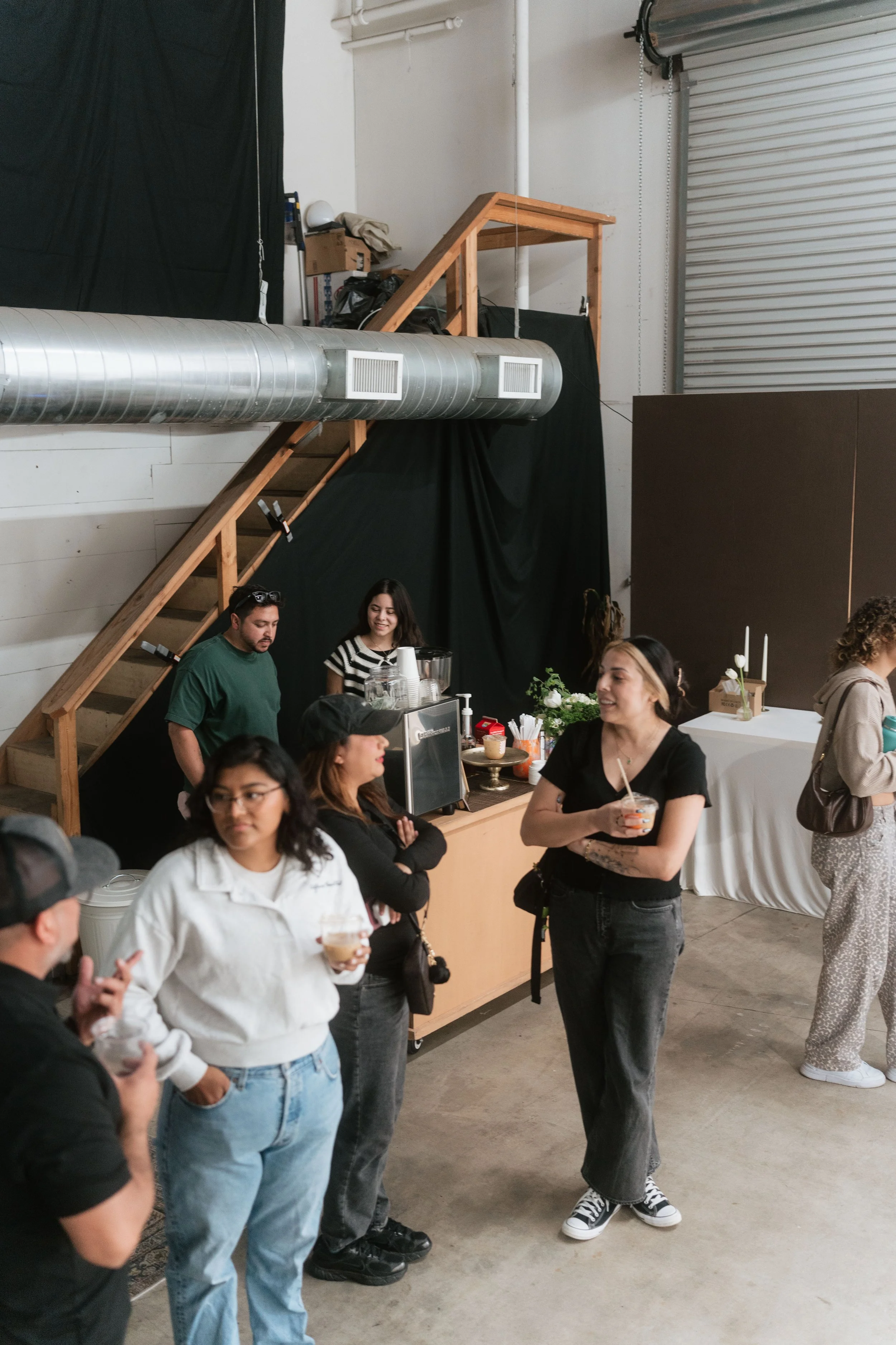 People standing and chatting in a room with a black backdrop, a staircase, and a table with flowers and drinks.