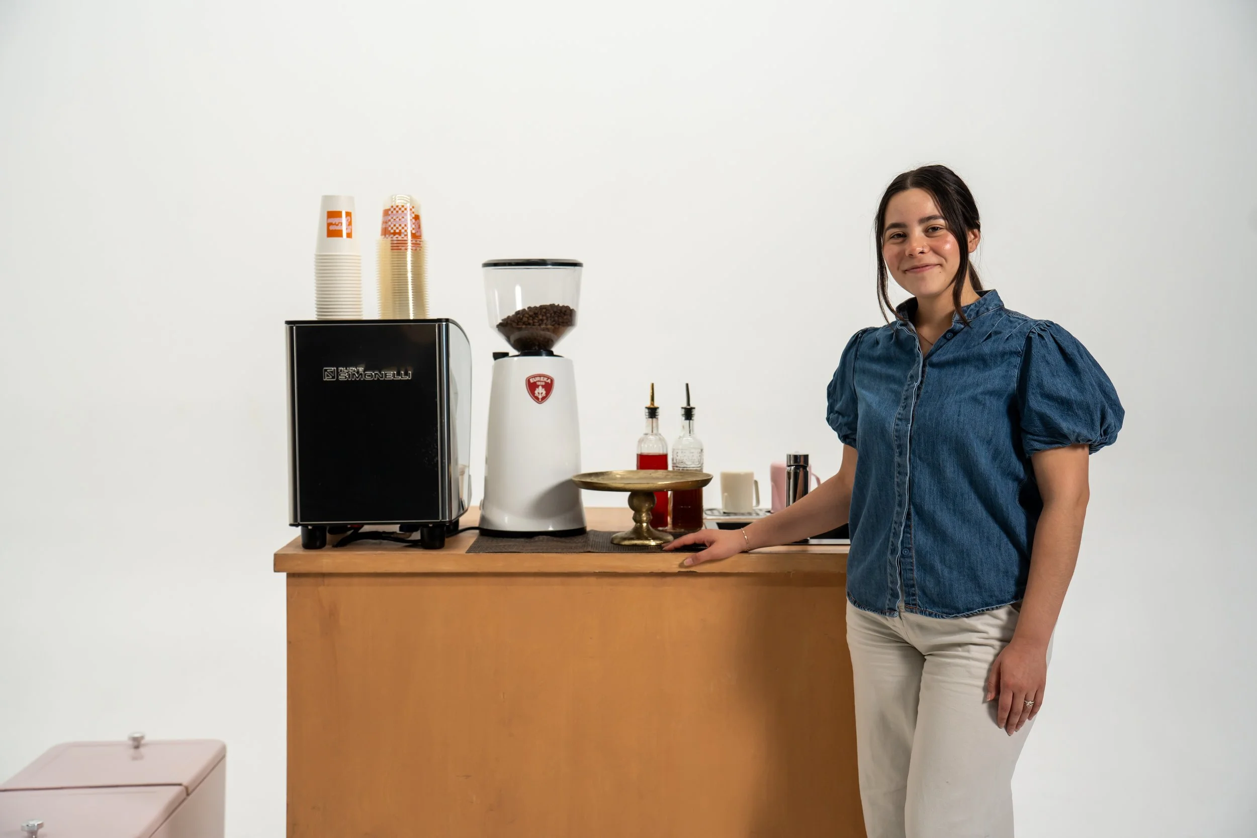 A woman stands behind a counter with coffee making equipment, including a espresso machine, grinder, and various syrups, in a minimalist setting with a white background.