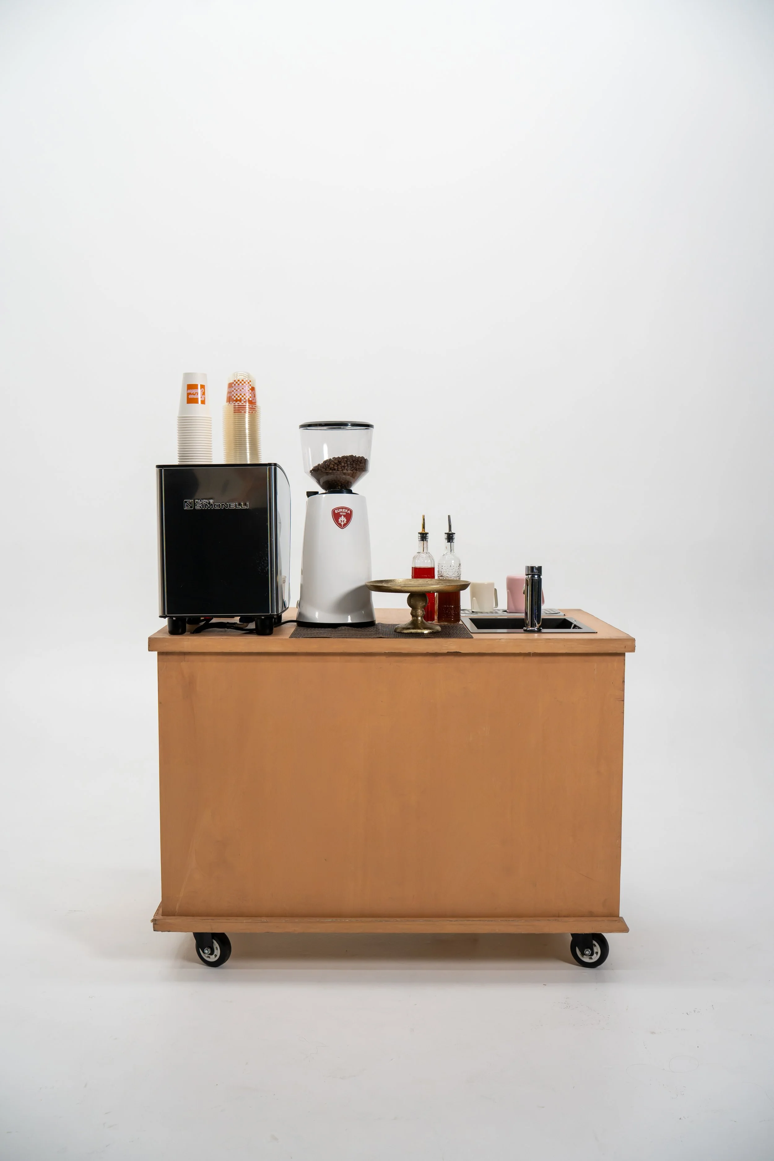 A mobile wooden coffee cart on wheels with a small sink and brewing equipment, including a coffee grinder, next to a plain white wall.